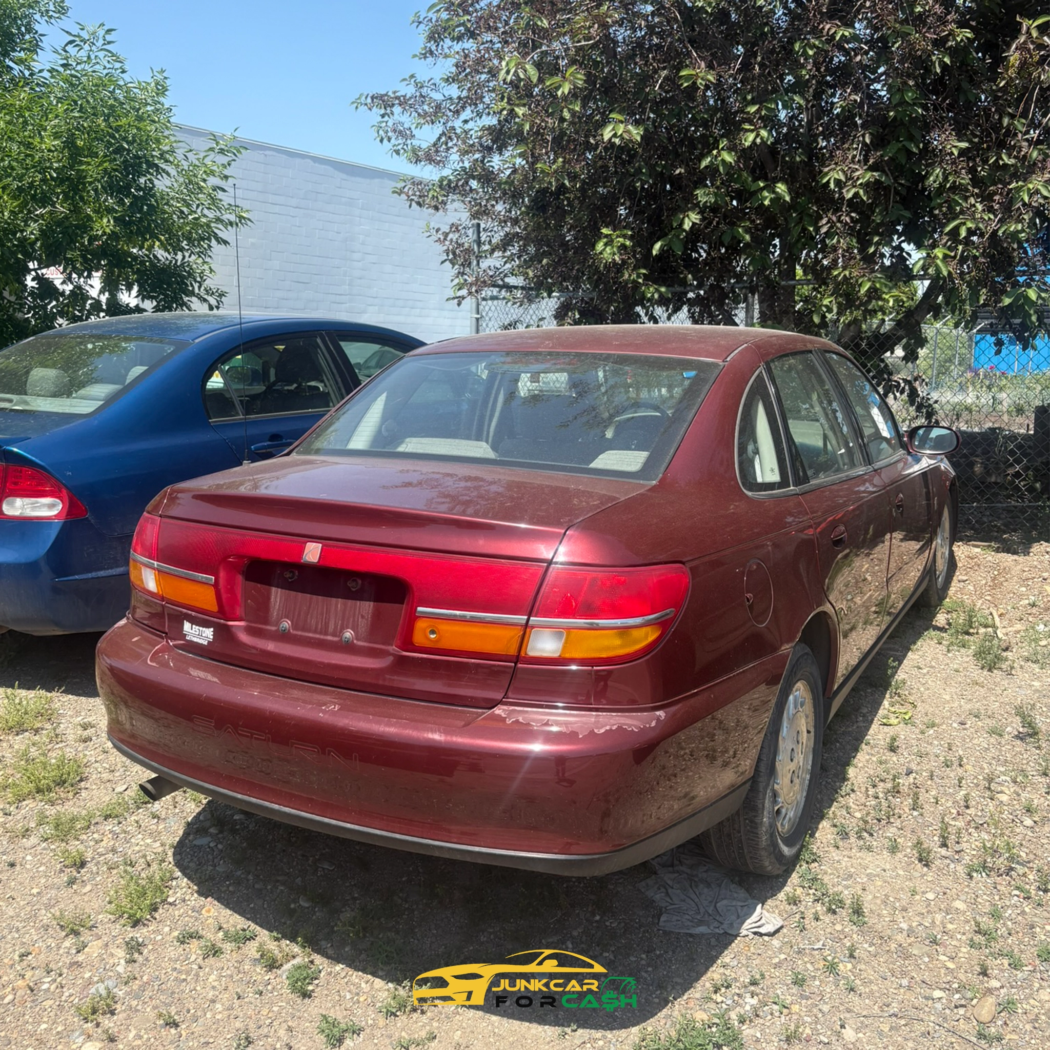 "Maroon sedan with missing rear plate and visible wear parked on dirt beside blue car near tree and gray building."