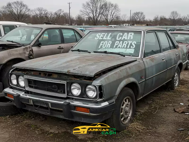 Rusty sedan with a "Sell Car for Parts" sign parked in a junkyard.