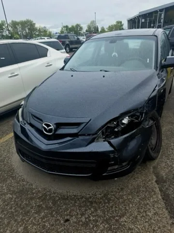 Dark-colored Mazda with front right damage including broken headlight, misaligned bumper, and oddly angled wheel; parked in lot with debris or fluid beneath damaged area.