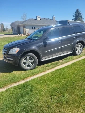 Black Mercedes-Benz GL-Class SUV parked with front wheels on sidewalk and rear on grass; sunny residential scene with trees and single-story house.