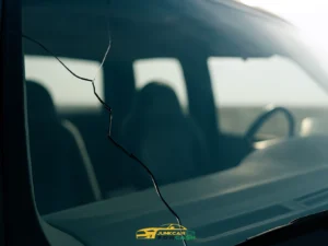 Front windshield of vehicle with long vertical crack extending diagonally across glass; interior visible with steering wheel and seats, sunlight streaming in from right side.