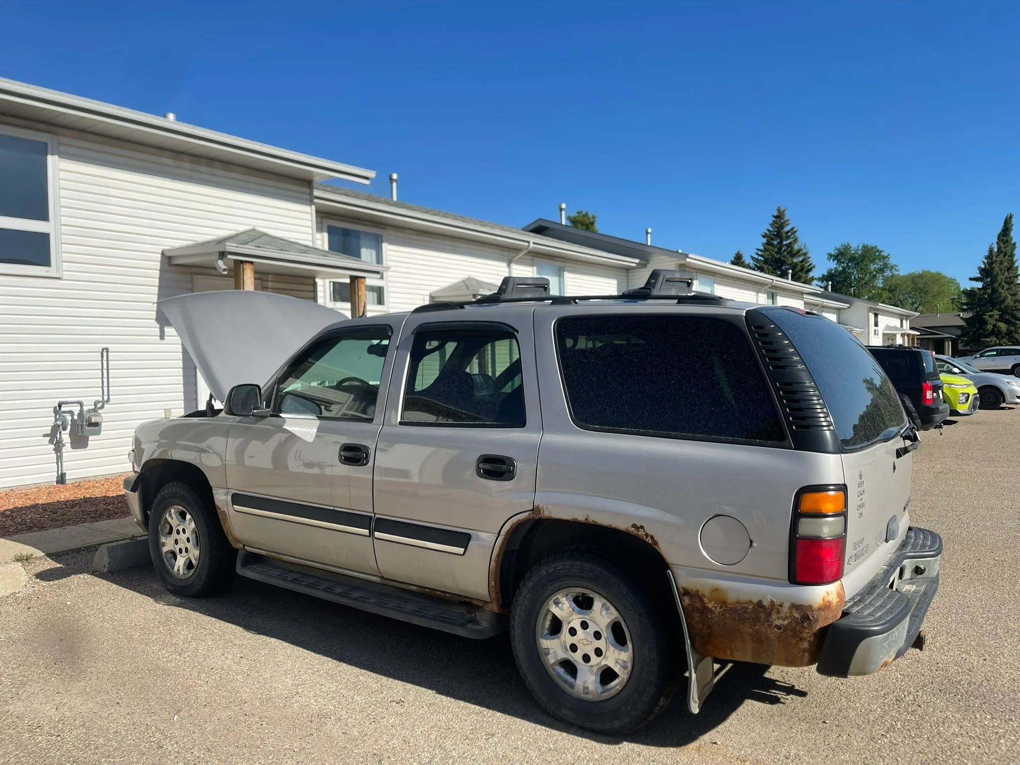 Silver SUV with rust damage and open hood parked near white multi-unit building; sunny day with clear sky and other vehicles nearby.
