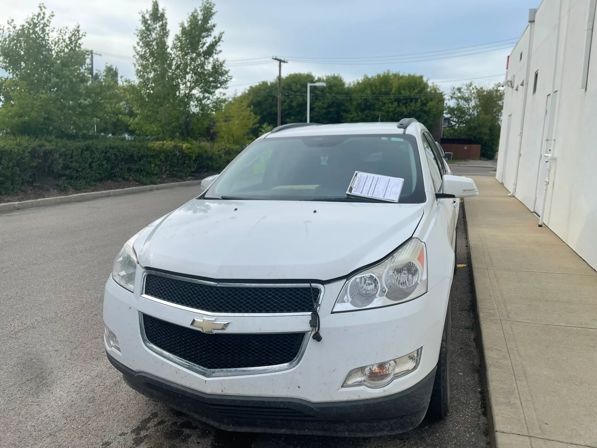 White Chevrolet SUV parked beside building with paper under windshield wiper; partly cloudy sky, sidewalk and greenery in background.