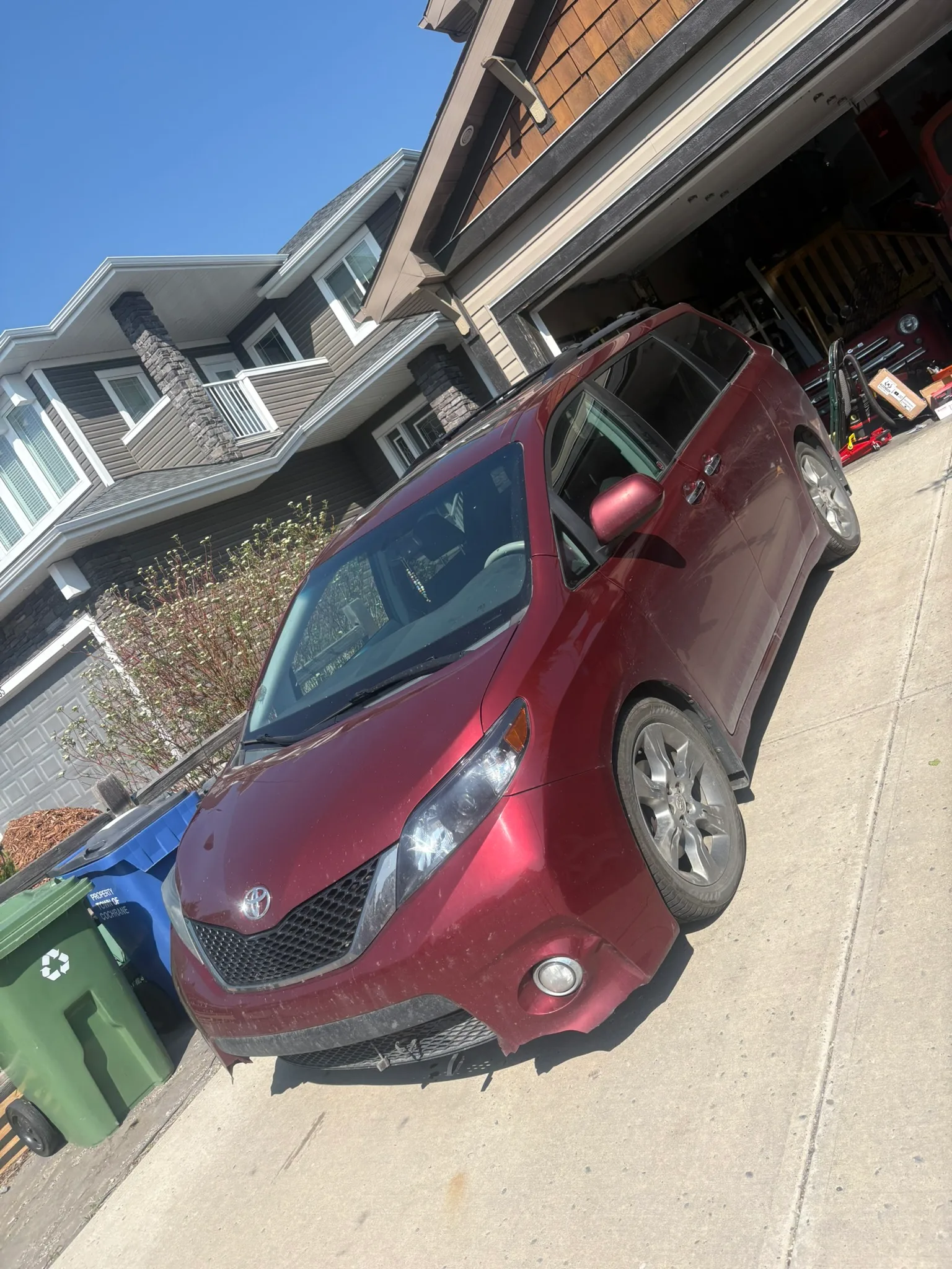 Red Toyota minivan parked in suburban driveway with open garage; recycling bins nearby, sunny day with clear blue sky.