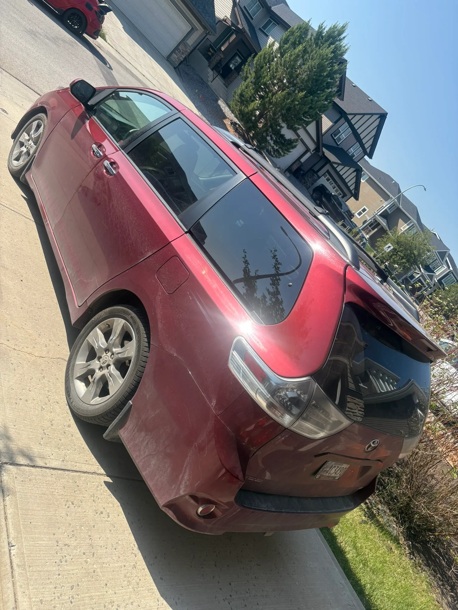 Maroon Toyota Sienna minivan parked on clean concrete driveway; light dust on lower panels, sunny suburban neighborhood with modern homes.