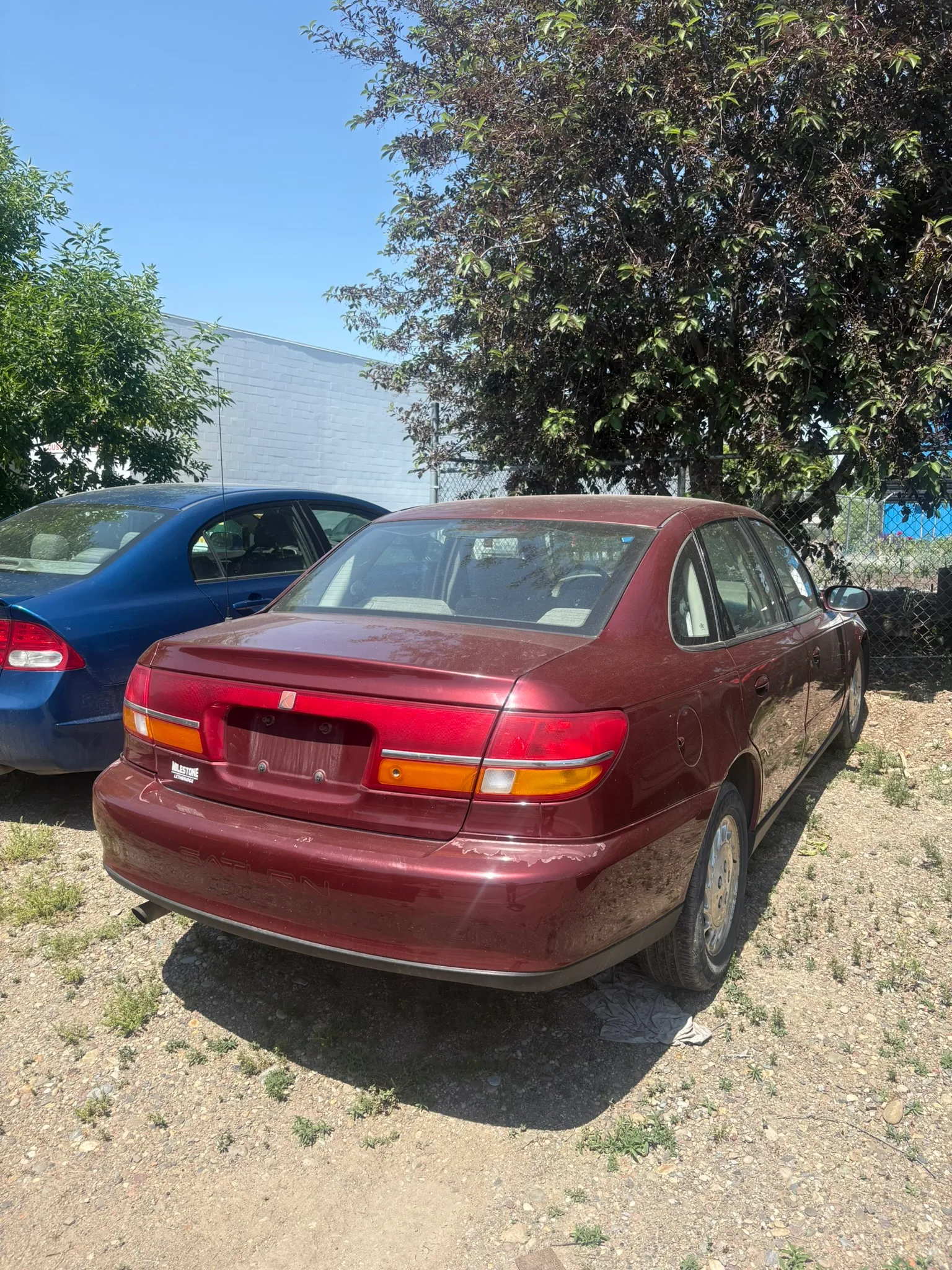 Maroon sedan missing rear plate parked on dirt lot beside blue car; older model with light grime, tree and gray building in background.