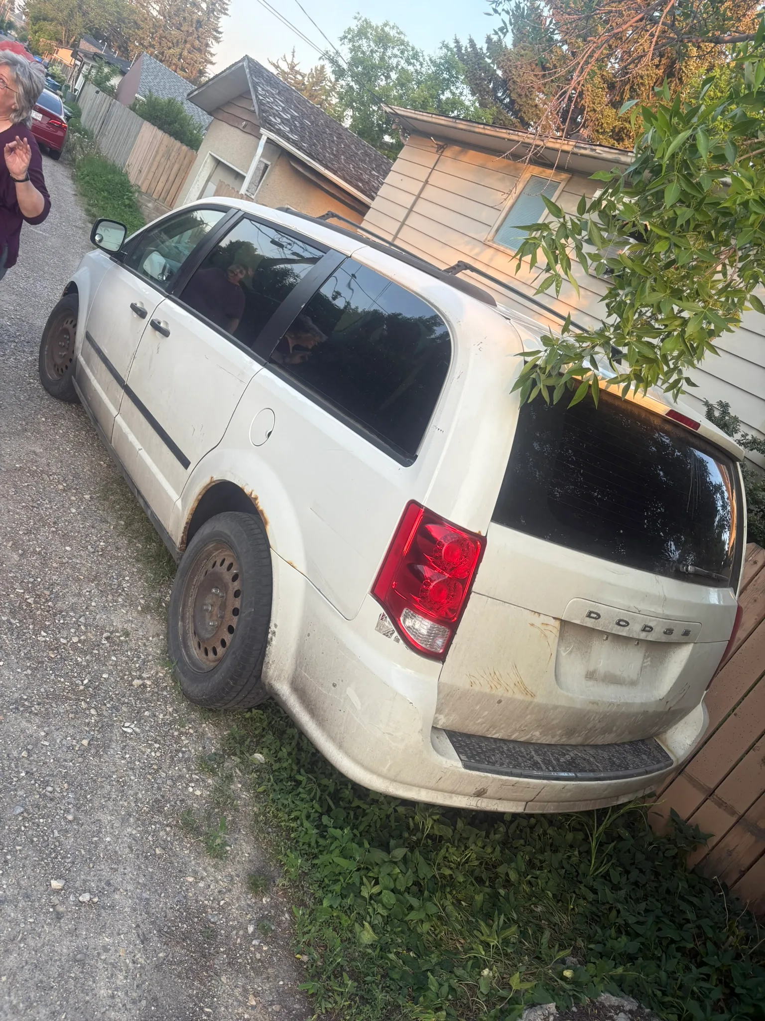 White Dodge minivan with rusted wheel wells and spare rear tire parked in gravel alley; wooden fence, house, and utility lines in background.