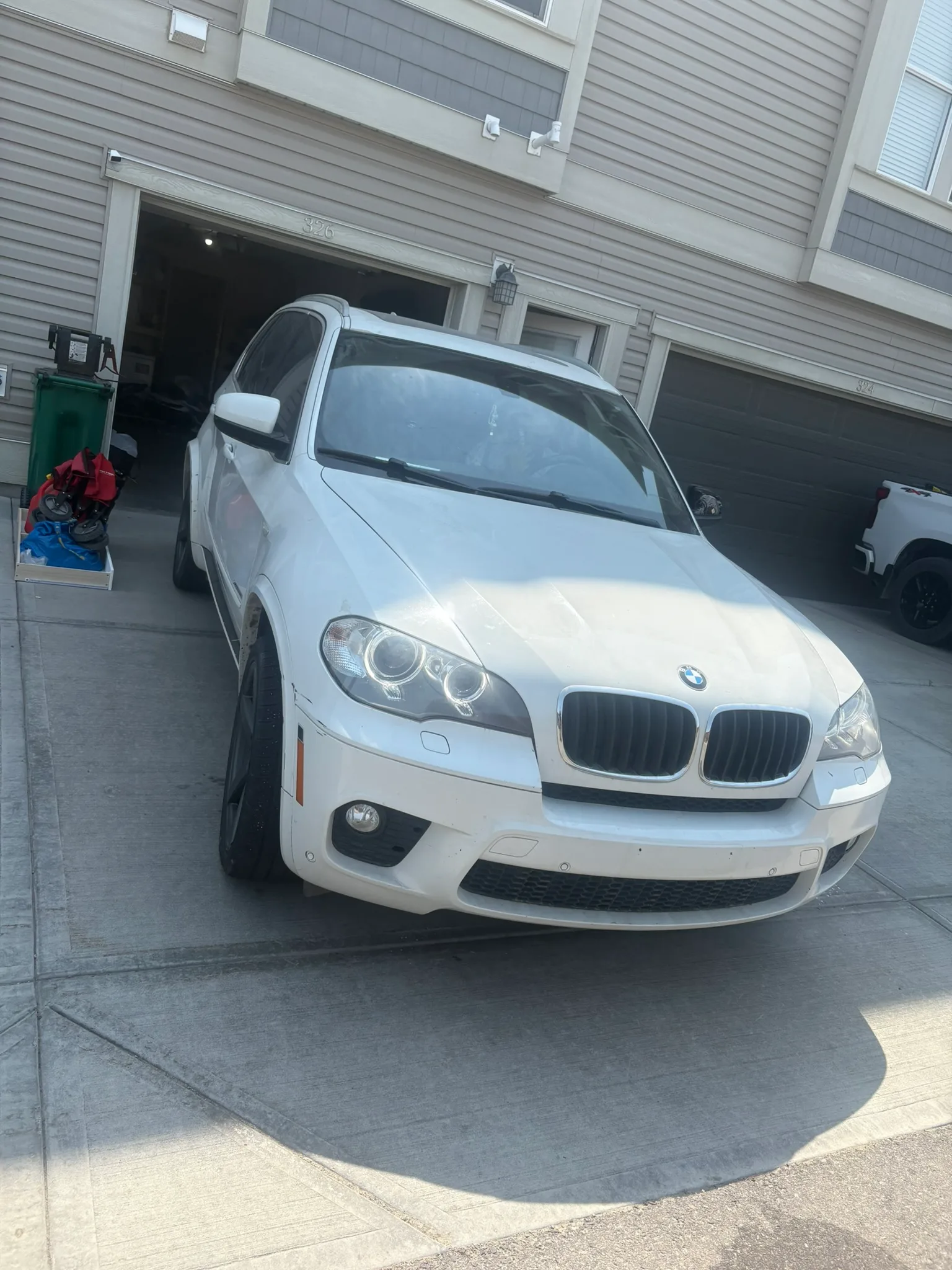 White BMW SUV with headlights on parked in townhouse driveway; clean exterior, beige siding and garage doors in background.