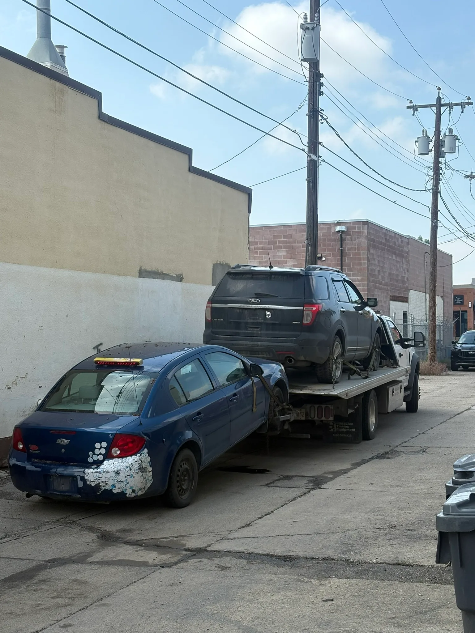 Tow truck hauling damaged black SUV with shattered rear window and blue sedan with dented bumper; alleyway lined with buildings and overhead wires.