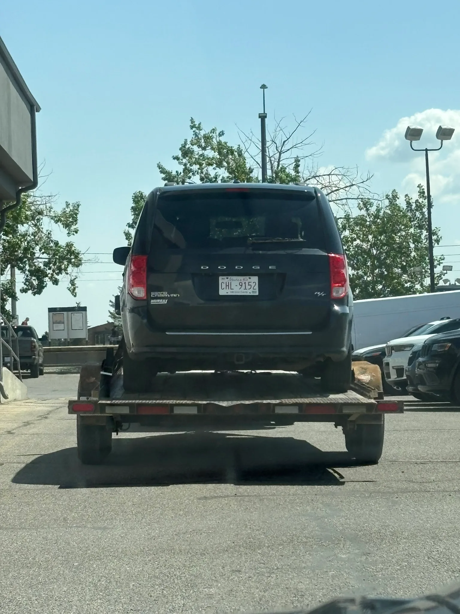 Black Dodge minivan with Alberta plate “CHL 9152” secured on flatbed trailer; parked in paved lot with trees, building, and clear sky in background.