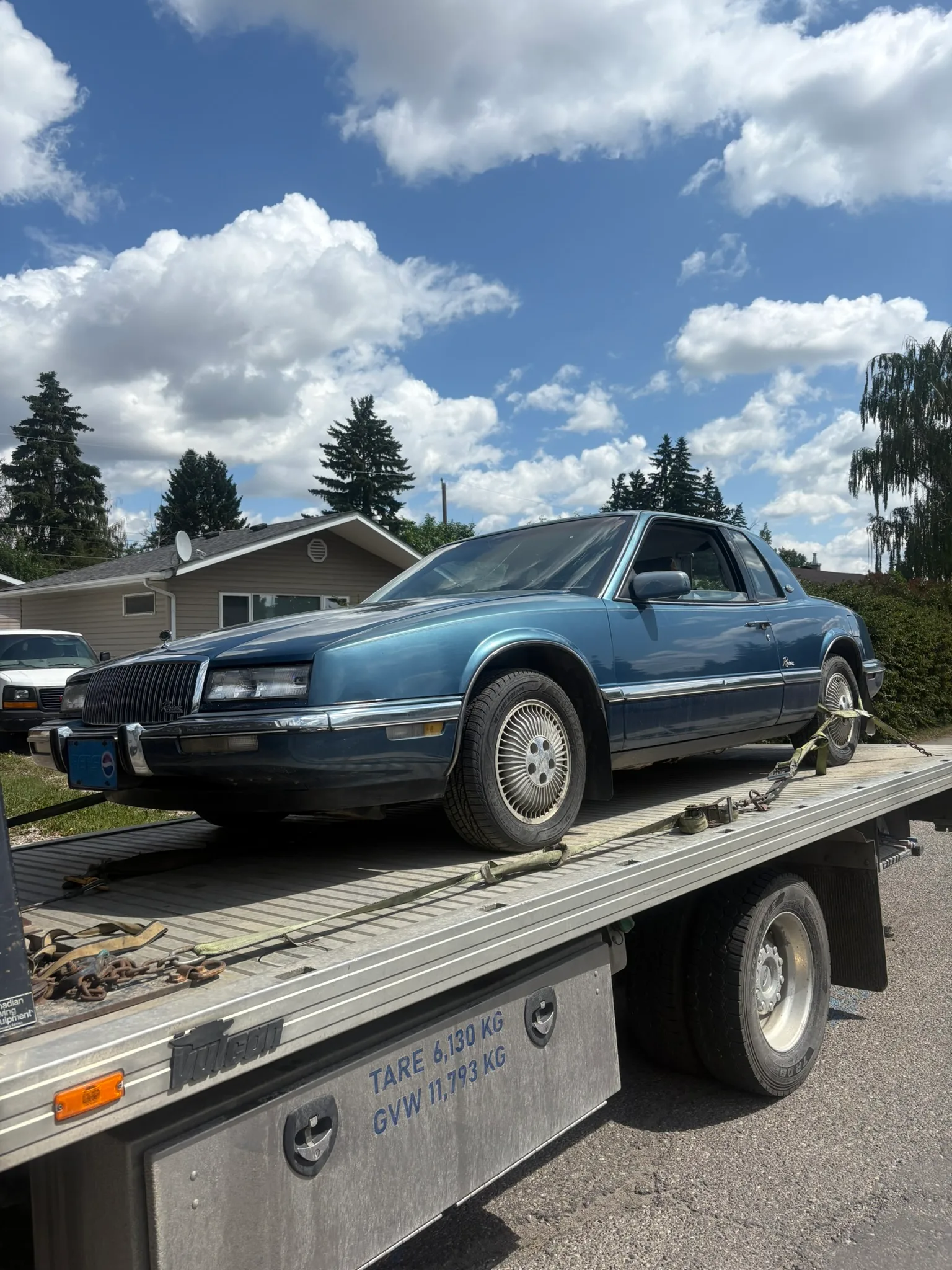 Blue two-door coupe secured on flatbed tow truck with visible weight markings; older model with straps on wheels, suburban homes and cloudy sky in background.