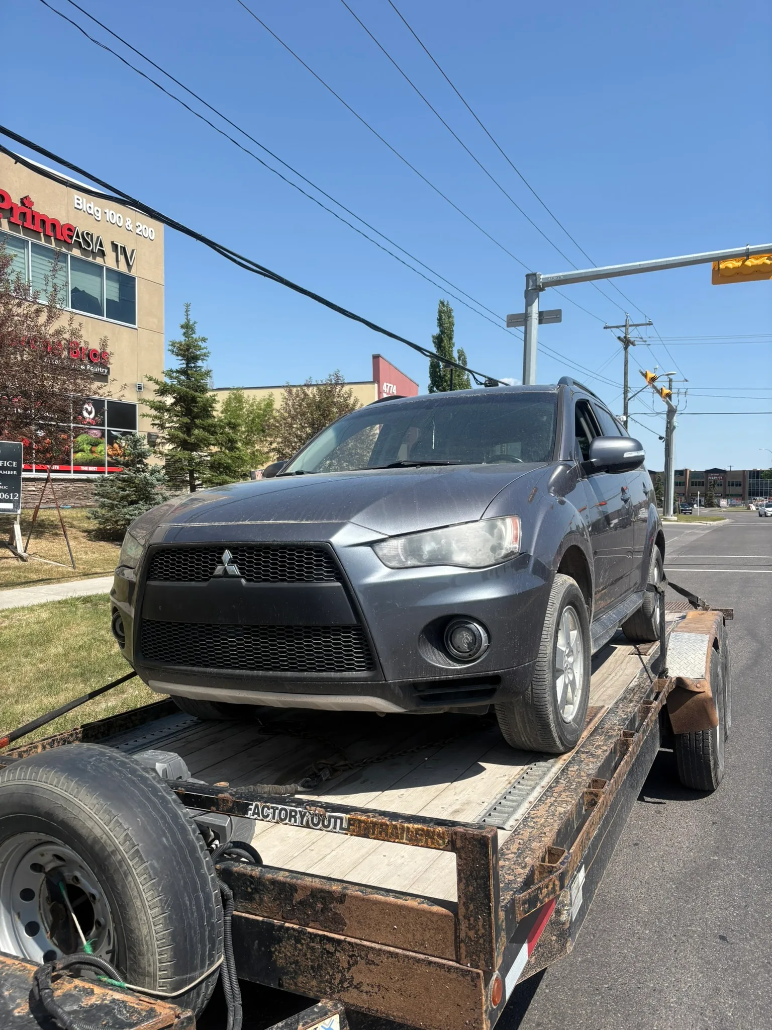 Dark gray Mitsubishi SUV secured on flatbed trailer in urban street; commercial signage, traffic light, and power lines visible under partly cloudy sky.