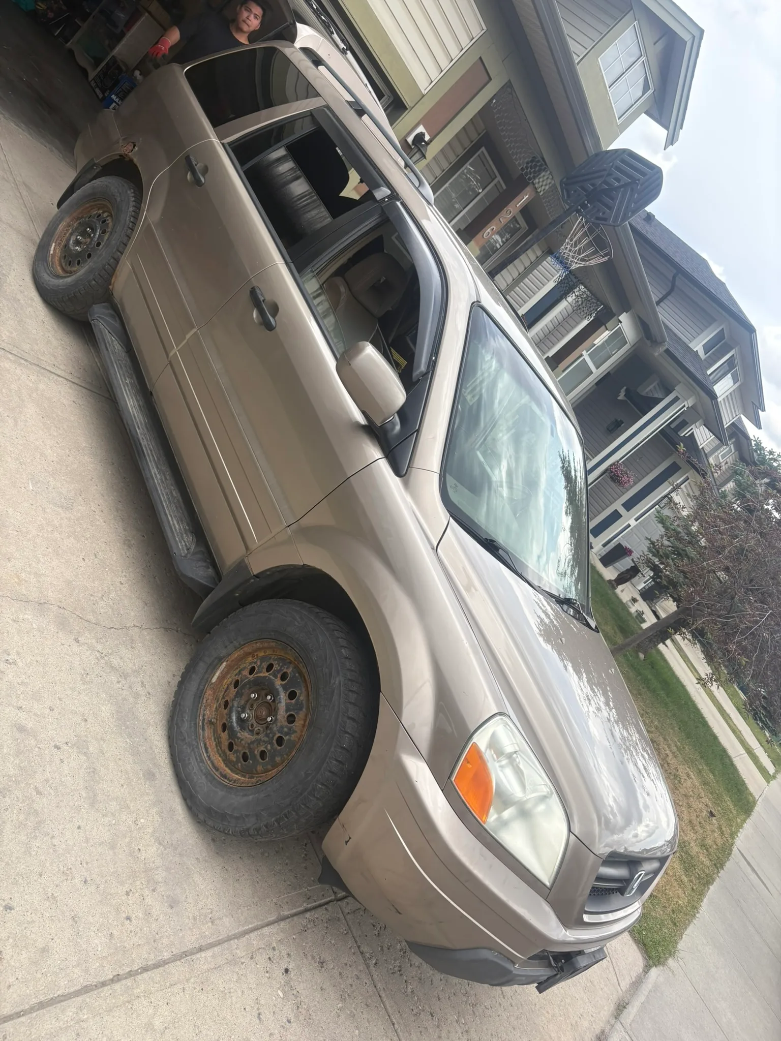 Beige SUV parked in suburban driveway with missing front hubcap and rust on wheel; rear window down, person partially visible inside.