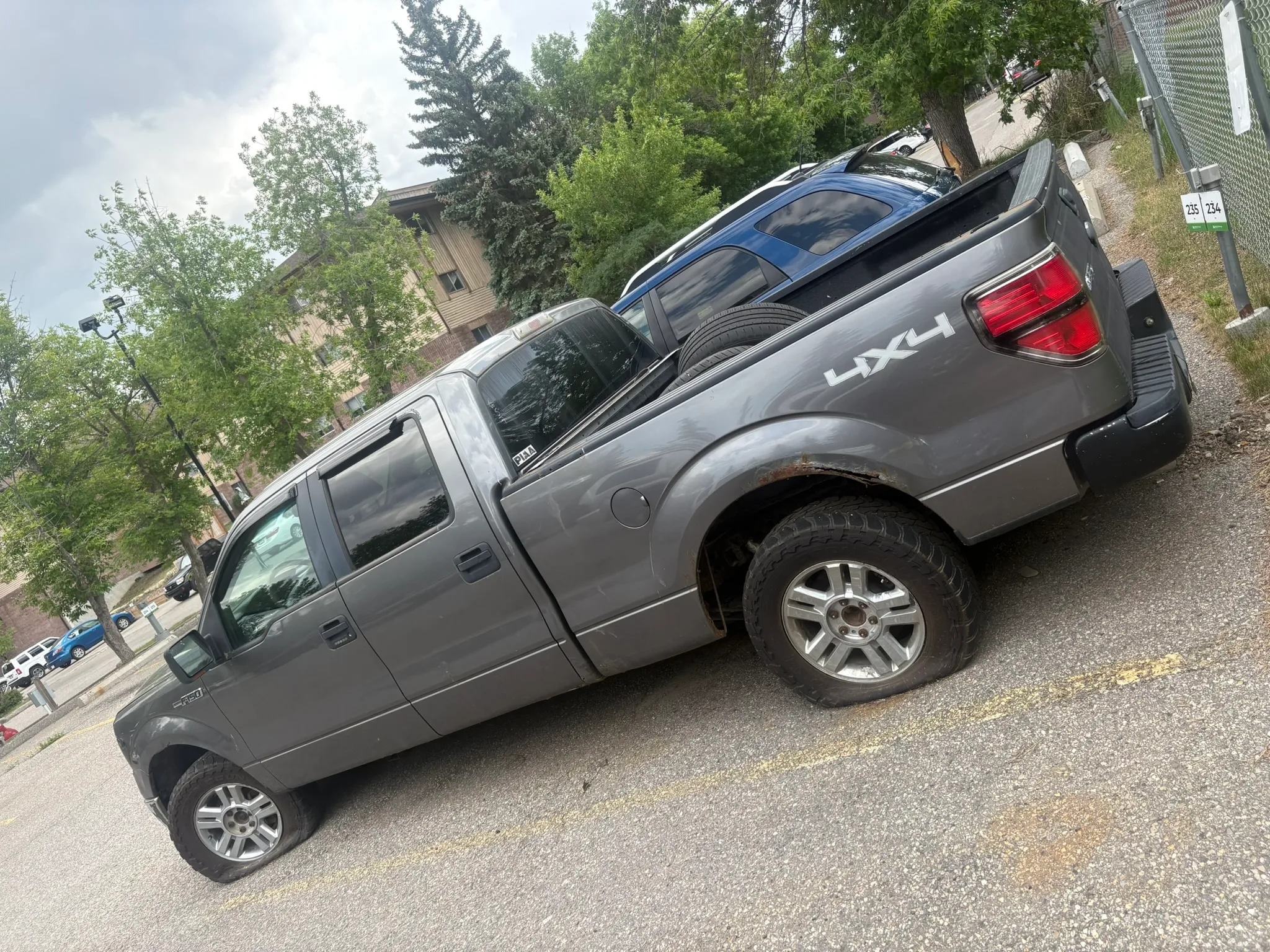 Gray Ford F-150 4X4 crew cab with visible rust on rear wheel well and spare tire in truck bed; parked beside blue SUV in lot with trees and building in background.