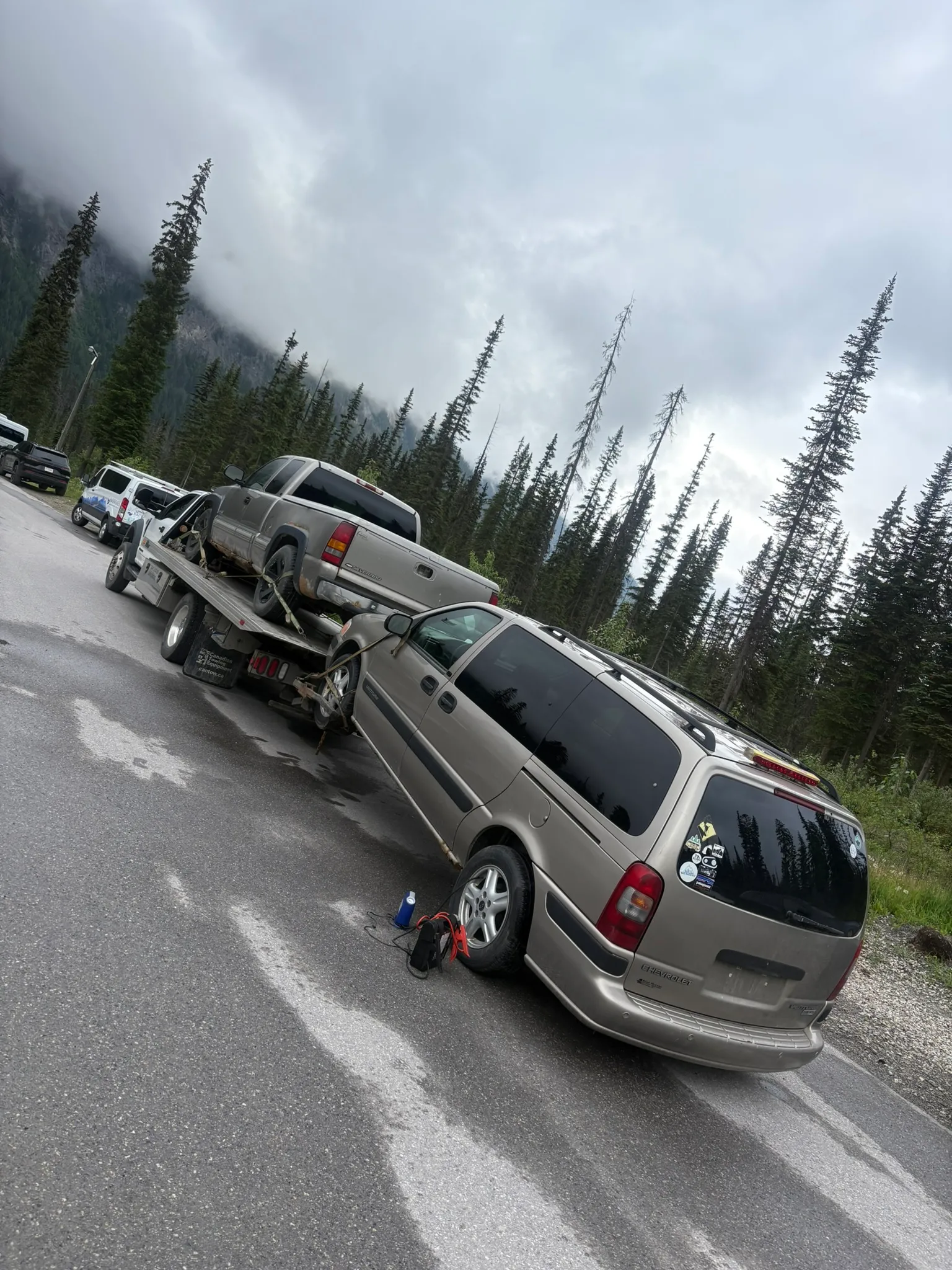 Tan SUV secured on tilted flatbed trailer towed by sticker-covered minivan; wet roadside with pine trees and mountains in background.