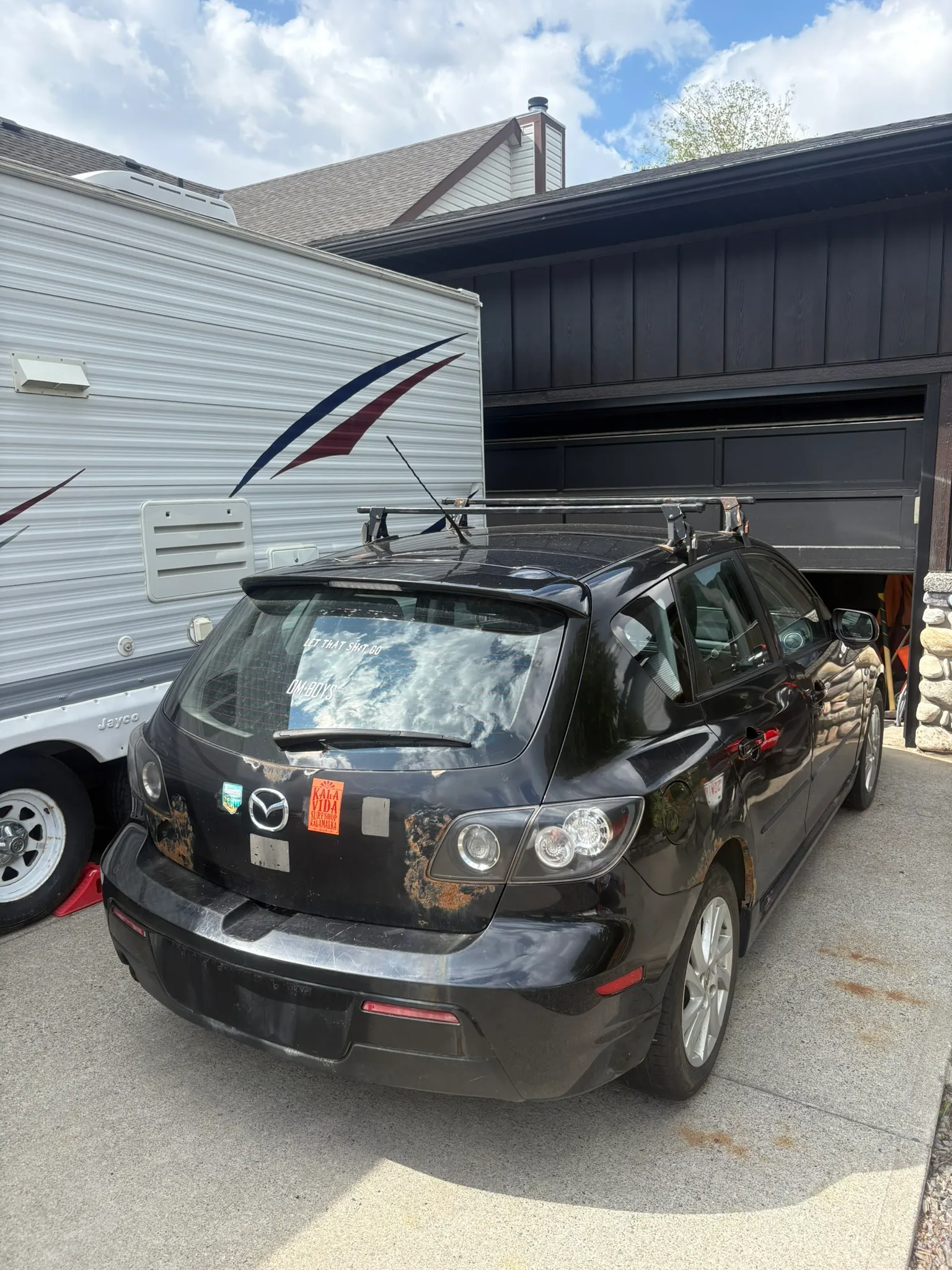 Black Mazda hatchback with rust near rear emblem and Yosemite sticker parked beside large white RV; stone-accented garage in background.