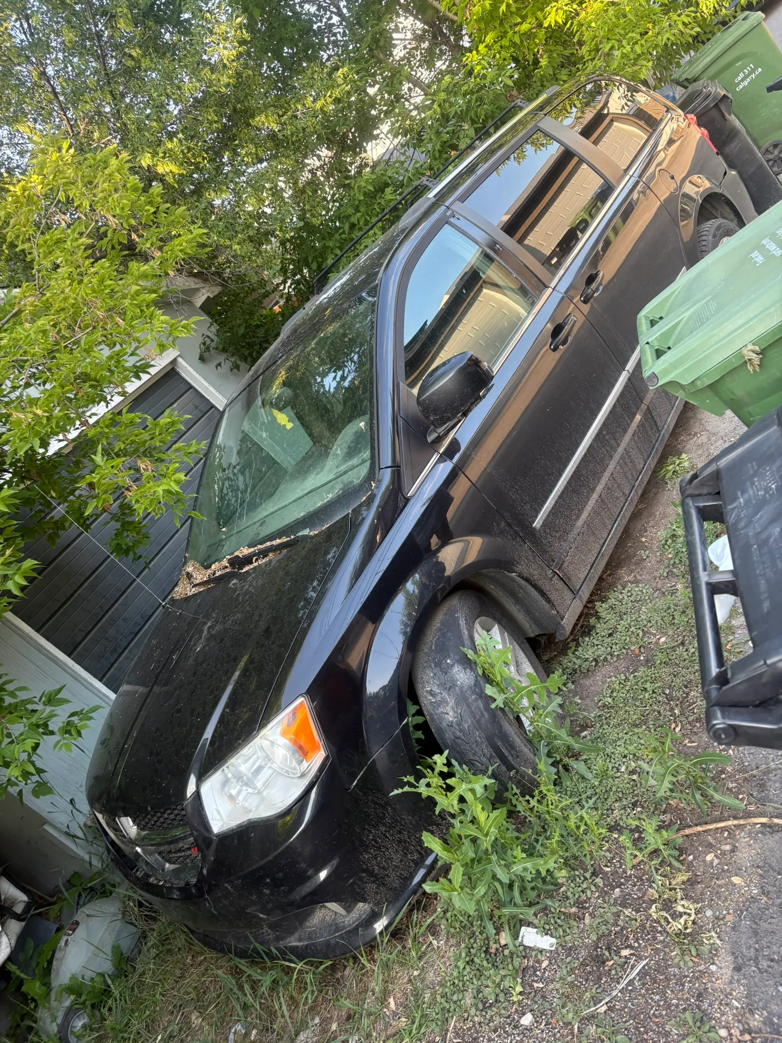 Black minivan with flat front tire and debris-covered windshield parked beside garage; surrounded by overgrown vegetation and trash bins on uneven surface.
