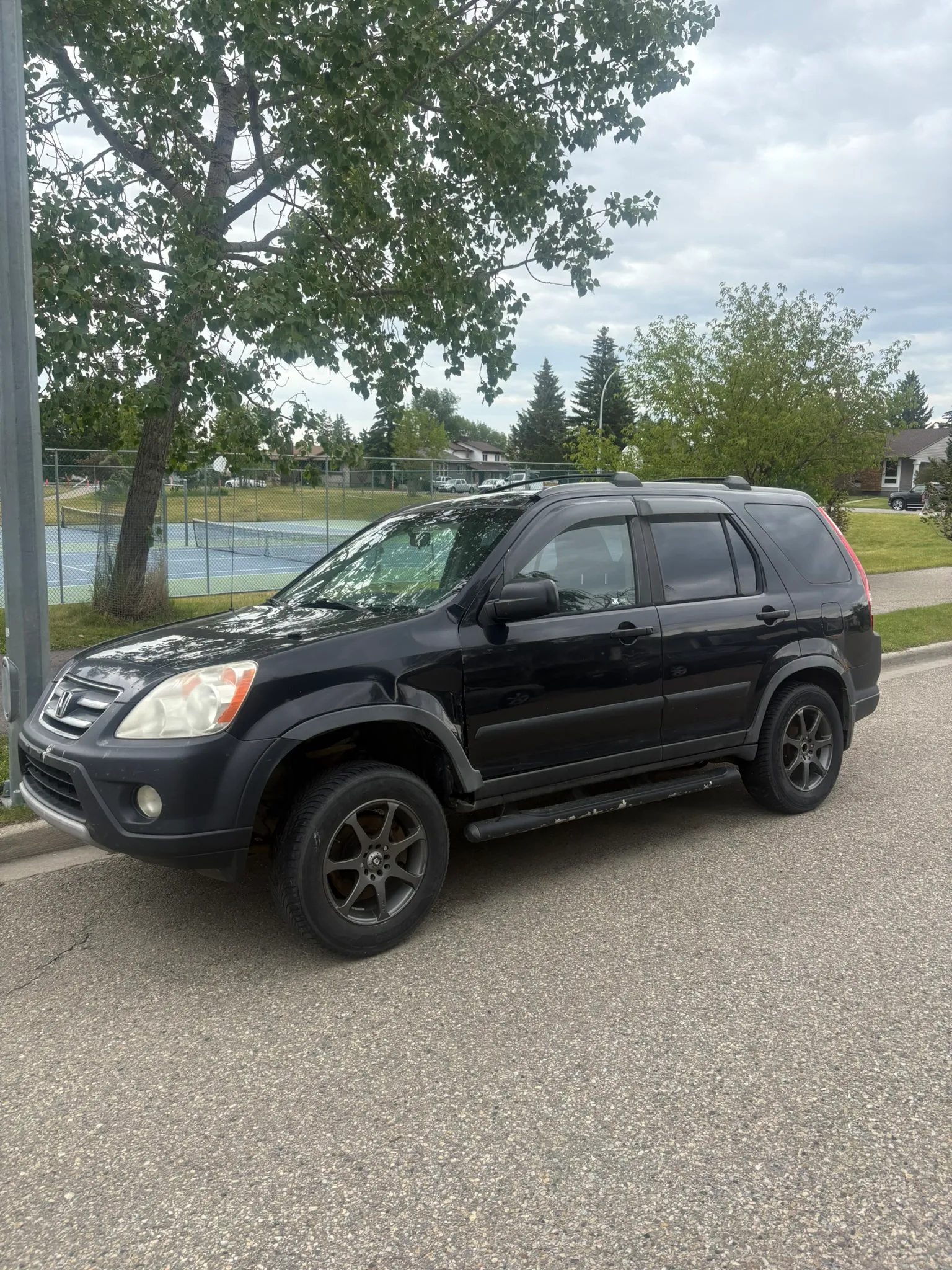 Black Honda CR-V with side steps and alloy wheels parked near tennis court; overcast sky, chain-link fence, and suburban homes in background.