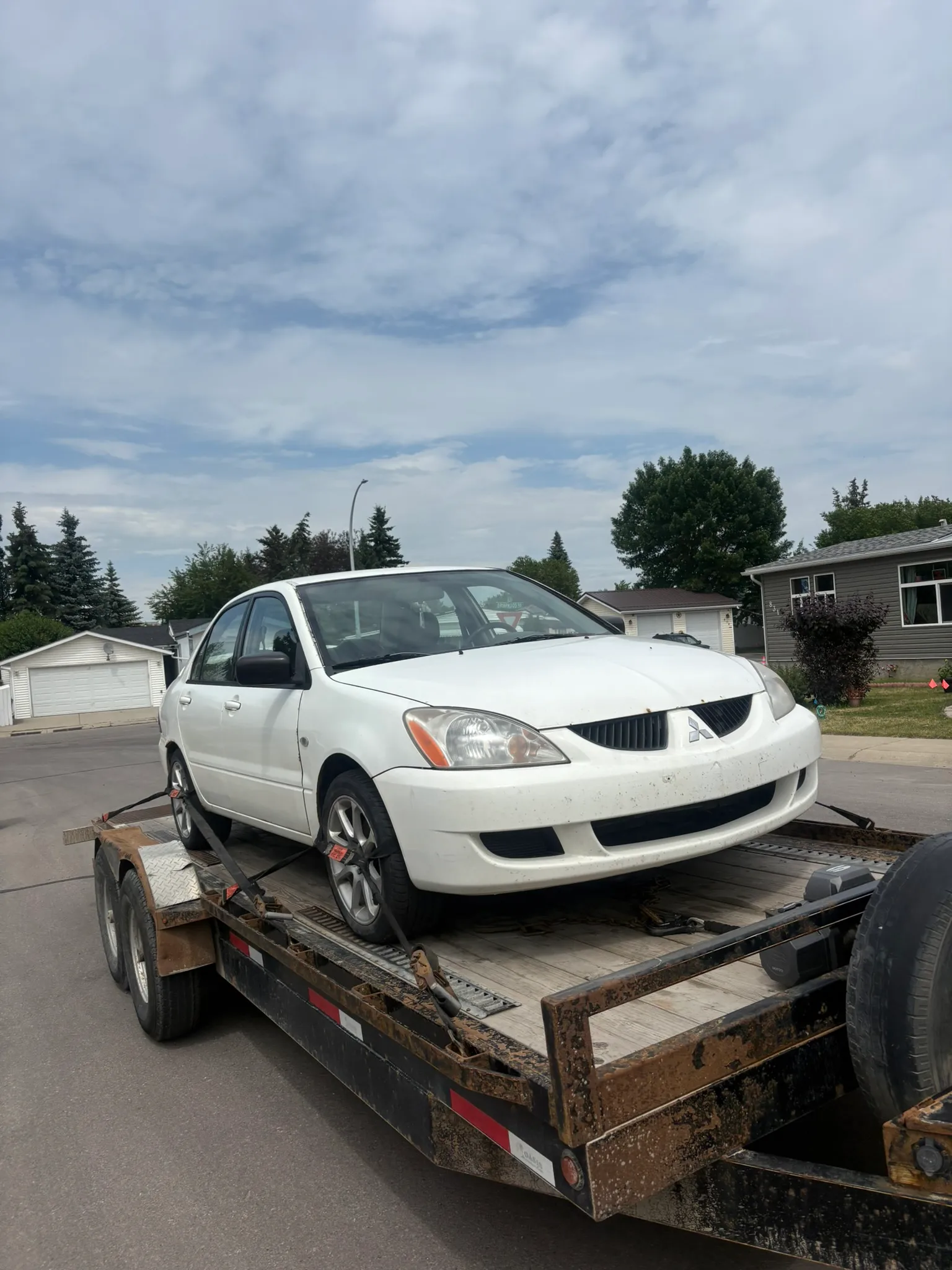 White Mitsubishi Lancer with missing front emblem secured on flatbed trailer; parked on residential street with cloudy sky and houses in background.