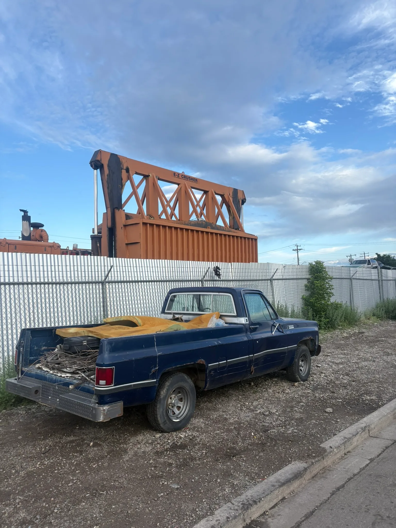 Weathered blue pickup truck with rust and debris-filled bed parked beside barbed-wire fence; industrial orange-and-black machinery visible in background under partly cloudy sky.