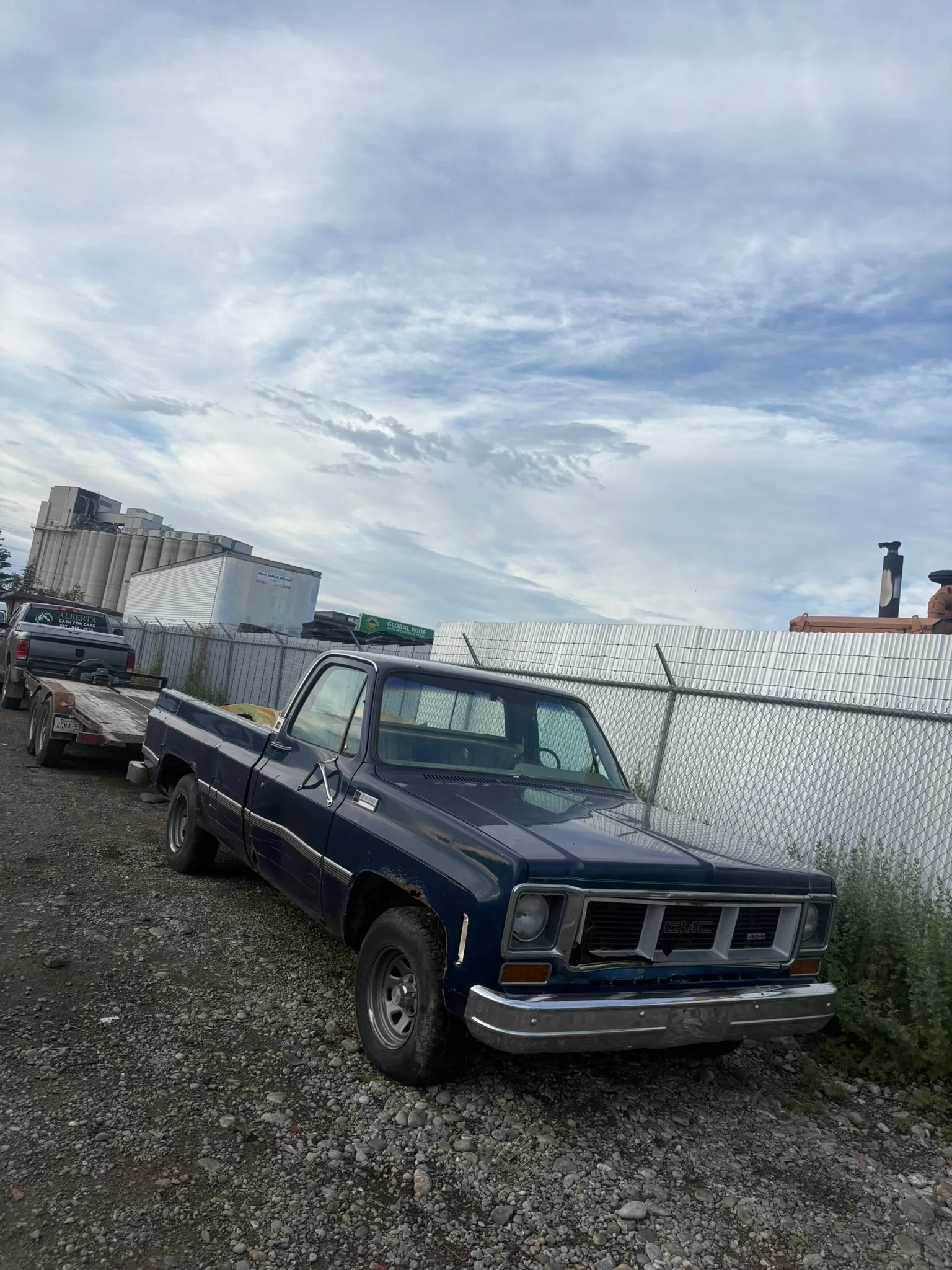 Old dark blue pickup truck with rust and missing grille parked on gravel near barbed-wire fence; industrial buildings and greenery in background under partly cloudy sky.