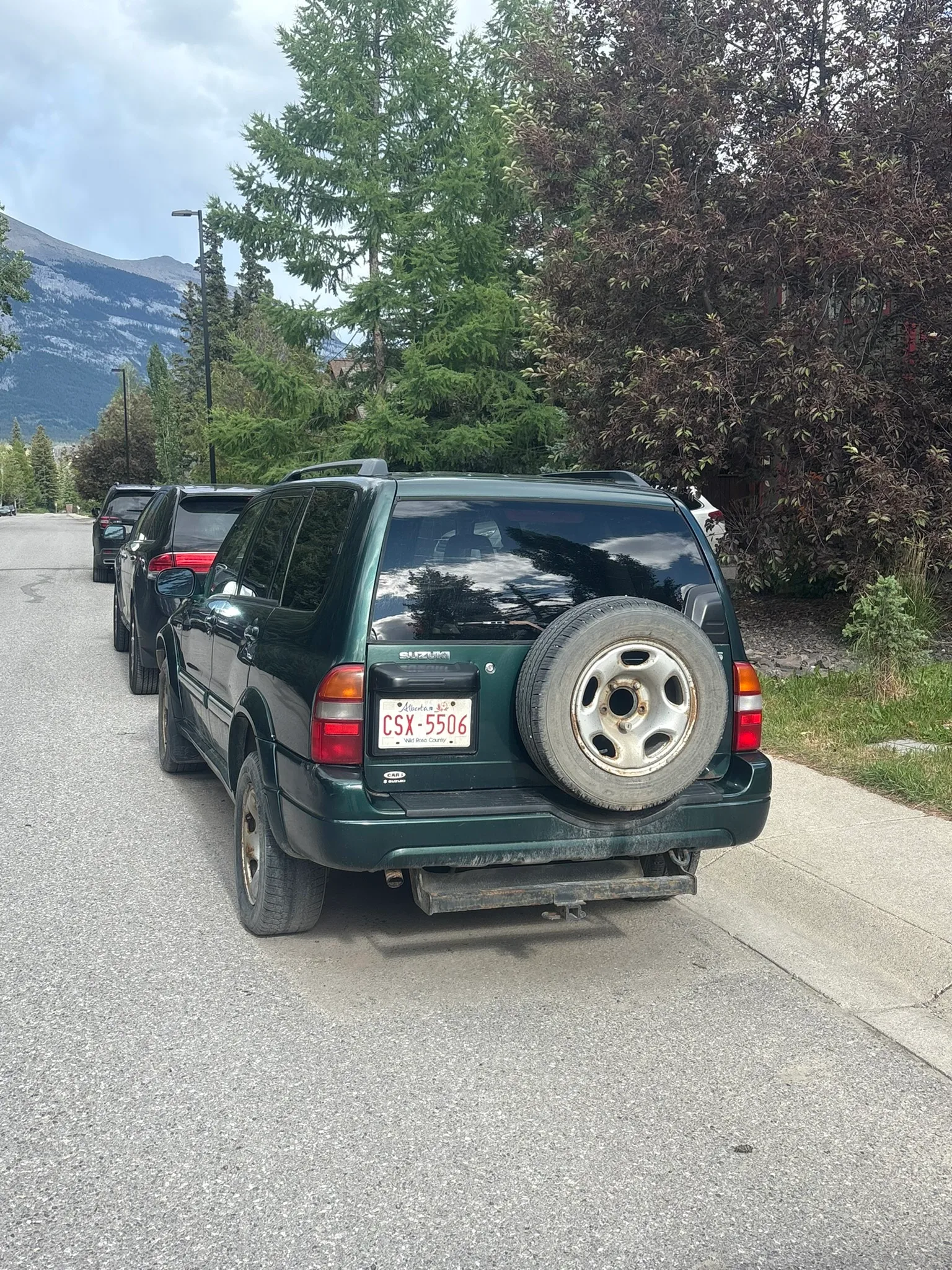 Green SUV with rear-mounted spare tire parked on residential street; Alberta license plate visible, mountains and trees in background.