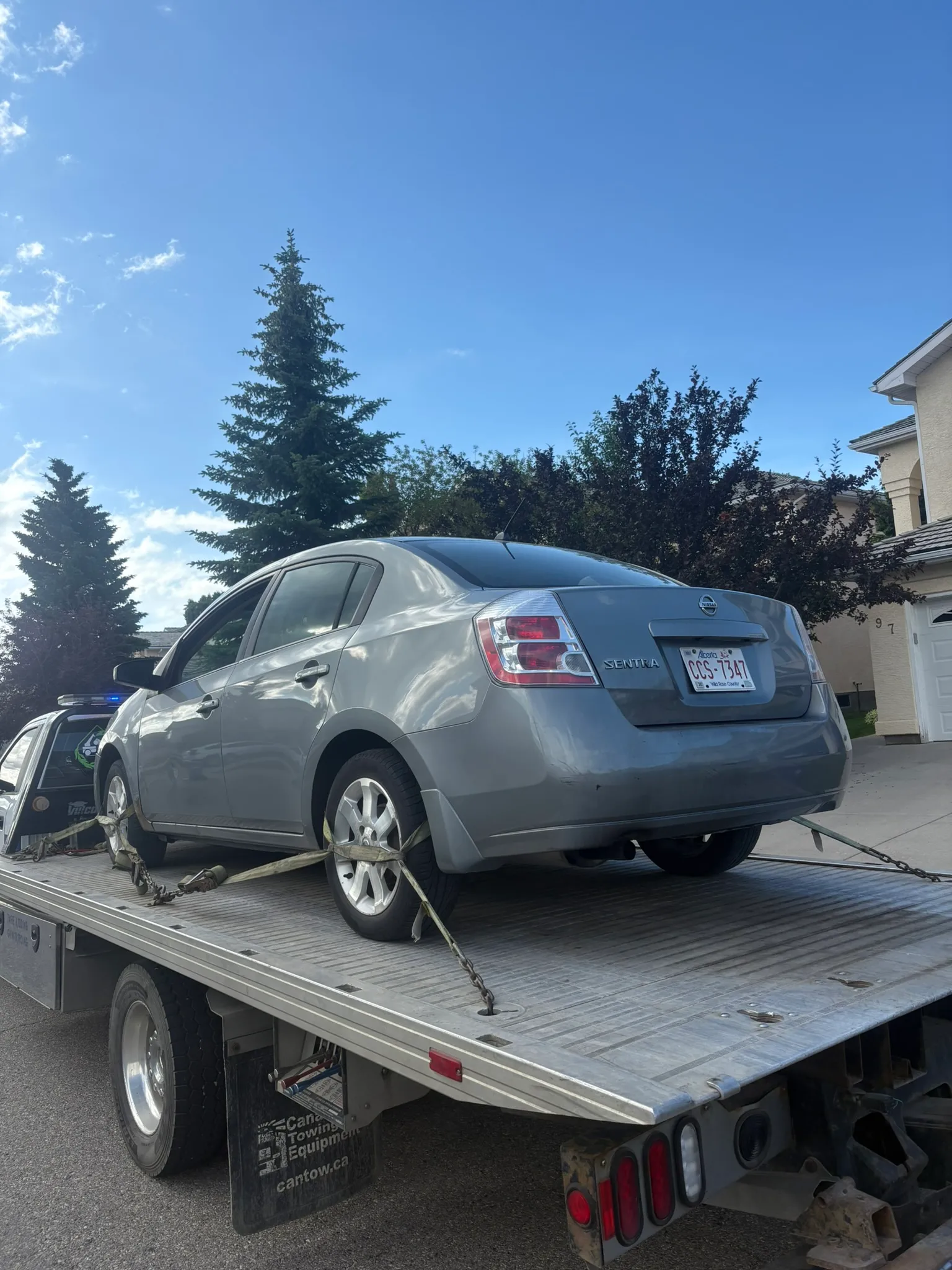 Gray Nissan Sentra with Texas plate secured on flatbed tow truck; parked on residential street with trees and houses under clear sky.