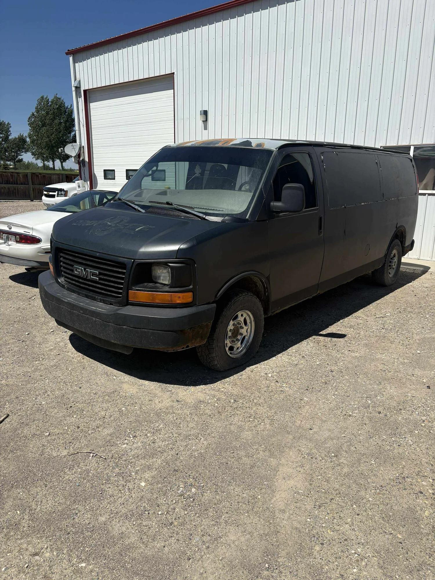 Matte black GMC Savana van parked outside white metal building with roll-up door; dusty lot and white car nearby suggest industrial setting.