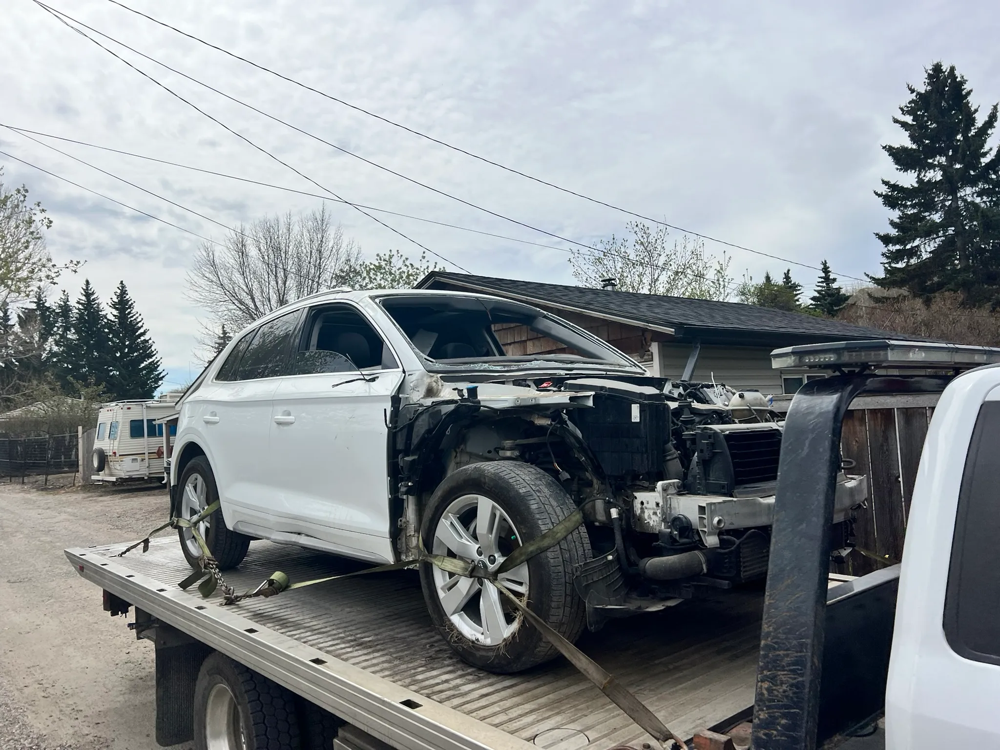 White SUV with missing front bumper and exposed engine area secured on flatbed tow truck; parked in suburban driveway with garage, trees, and RV in background.