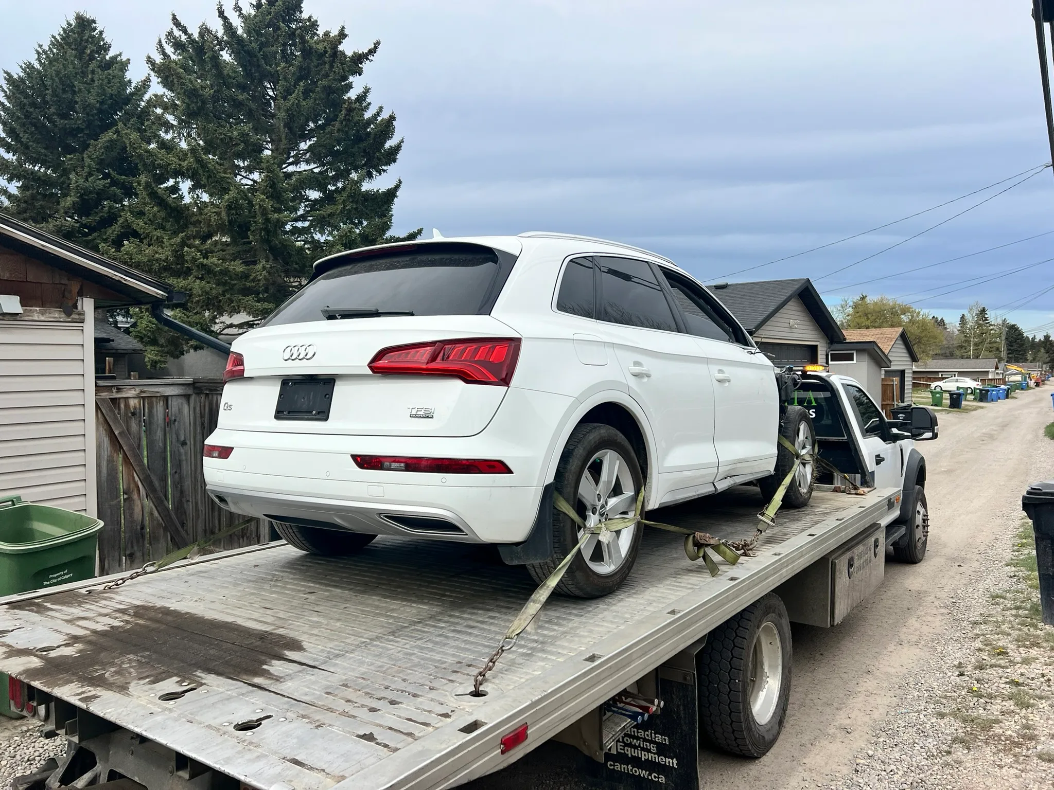 White Audi SUV in good condition secured on flatbed tow truck in residential alley; surrounded by houses, trees, and garbage bins under partly cloudy sky.