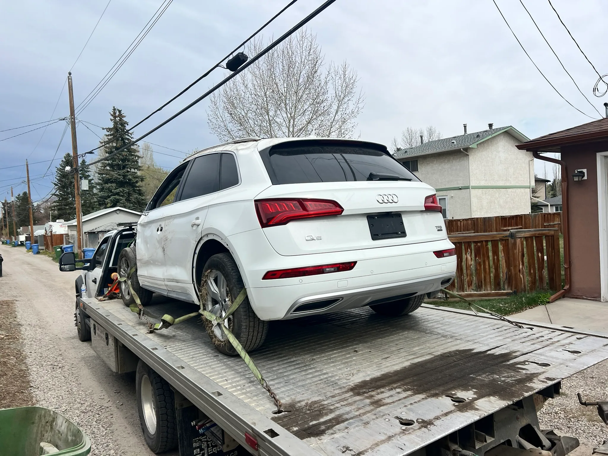 White Audi Q5 SUV in good condition secured with green straps on flatbed tow truck; parked in residential alley with houses, trees, and utility poles in background.