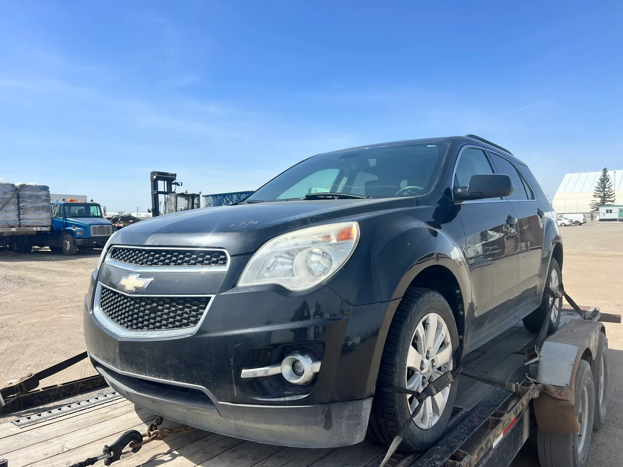 Black Chevrolet Equinox SUV in good condition secured on trailer in industrial lot; surrounded by blue truck, forklift, stacked materials, and white tent structure.