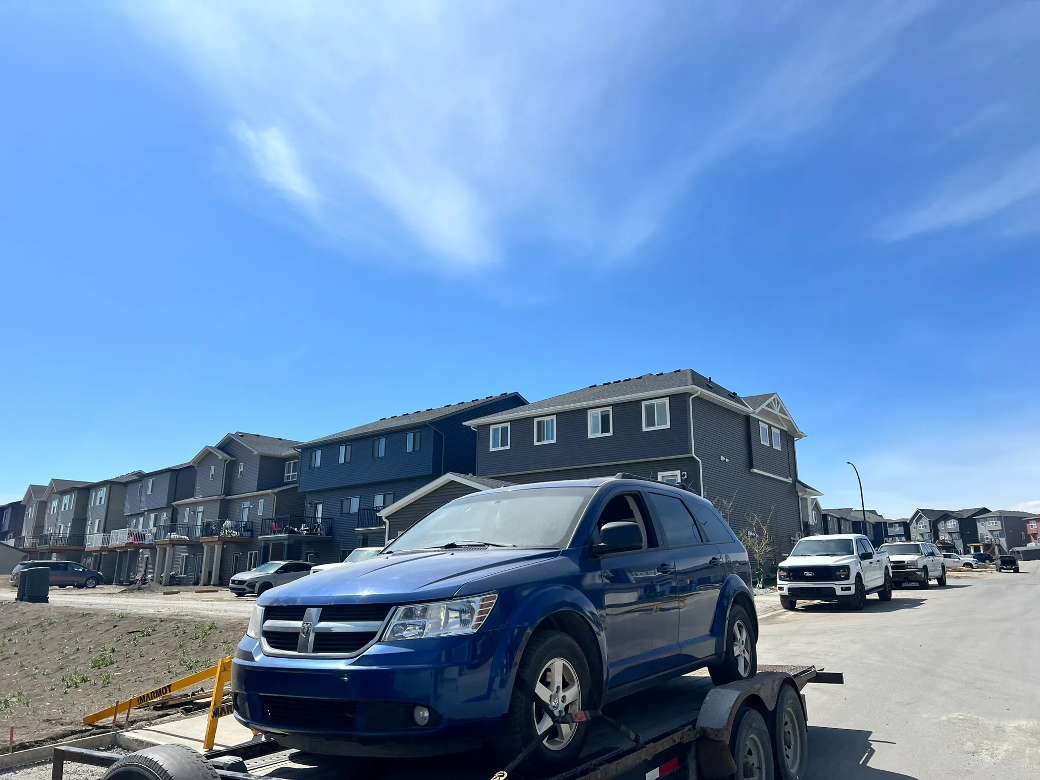 Blue Dodge Journey SUV secured on trailer hitched to white pickup truck; parked on residential street with modern townhouses and clear sky in background.