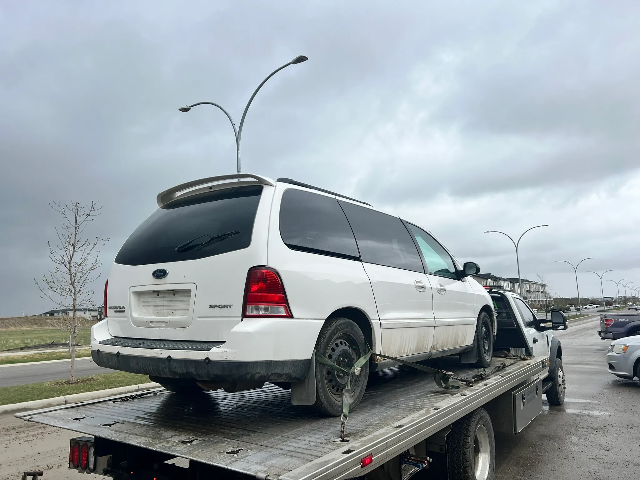 White Ford minivan with missing rear plate and spare rear left tire secured on flatbed tow truck; parked on street with buildings, vehicles, and cloudy sky in background.
