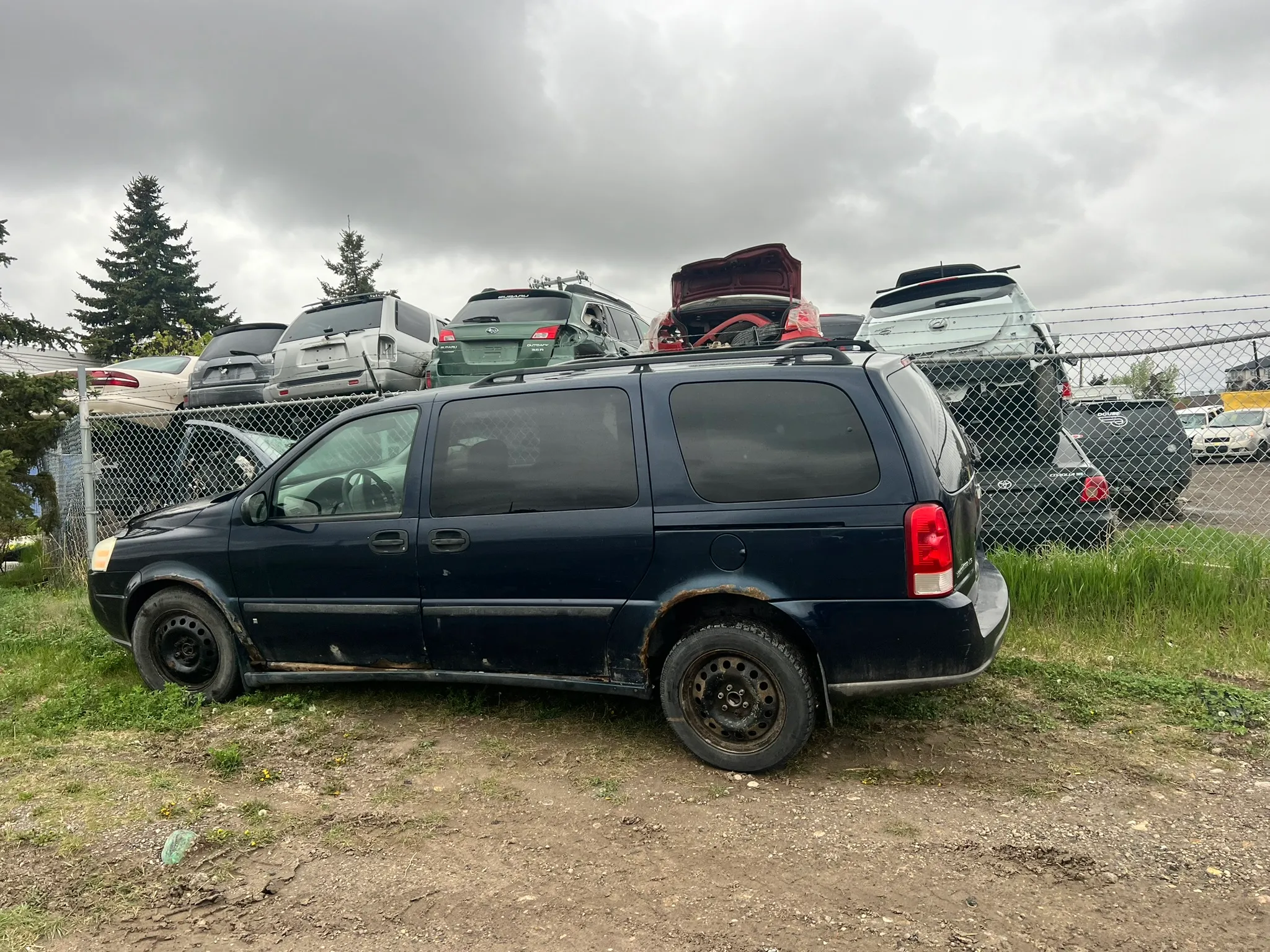 Dark blue minivan with rust and missing front hubcap parked on dirt near chain-link fence; stacked vehicles and overcast sky suggest salvage yard setting.