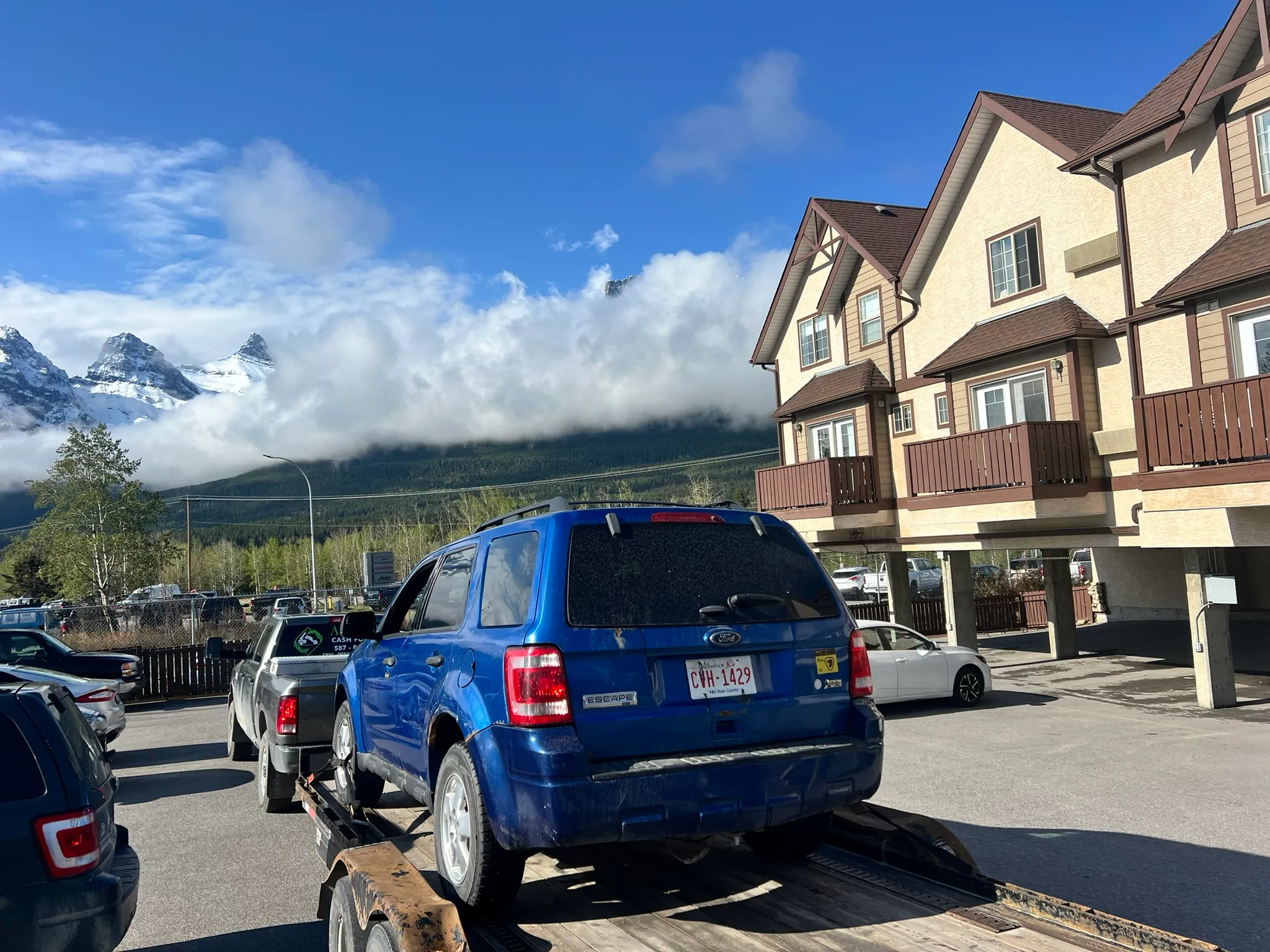 Blue Ford SUV with license plate CH-128P secured on trailer in residential lot; snow-capped mountains, forested base, and scattered clouds in scenic background.