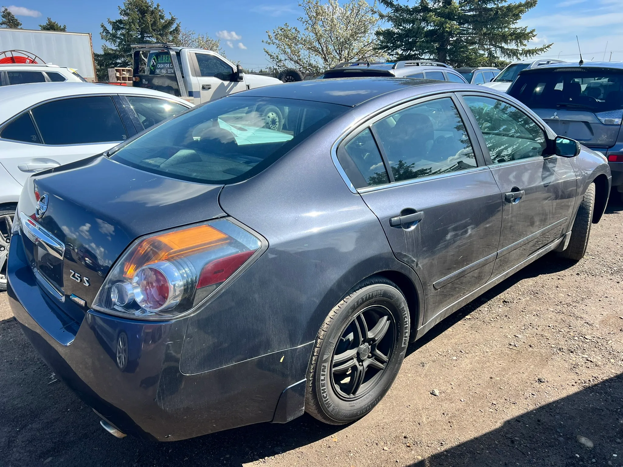 Dark gray Nissan Altima 2.5 S with black wheels parked in gravel lot among other vehicles; rear-left view shows taillights and used condition.