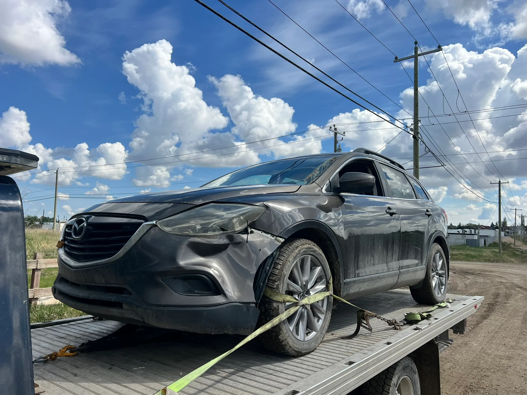 Damaged dark-colored Mazda sedan with crumpled front fender and green tie-down strap on flatbed tow truck; dirt road, utility poles, and partly cloudy sky in background.