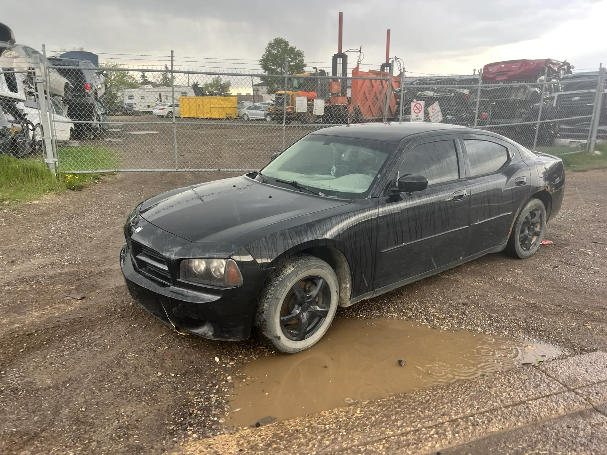Black Dodge Charger with mud-splattered body and front tire in puddle parked on gravel near chain-link fence; industrial machinery and trucks in background under overcast sky.