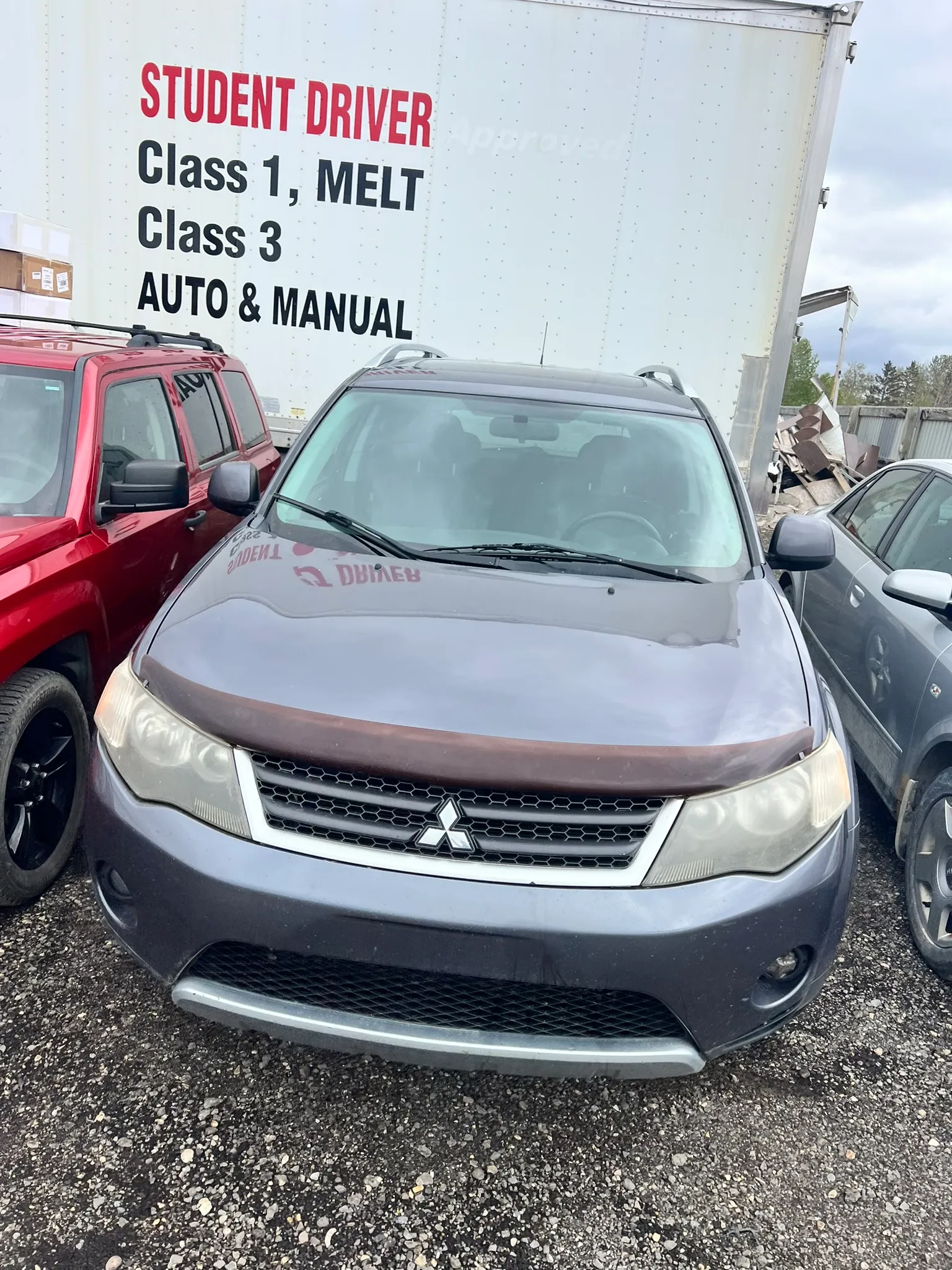 Grey Mitsubishi SUV parked between red SUV and grey sedan in lot; white trailer behind displays “STUDENT DRIVER Class 1, MELT Class 3 AUTO & MANUAL” text.