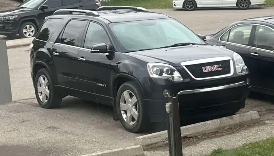 Black GMC Acadia SUV with chrome grille and wheels parked in outdoor lot beside other vehicles; grassy edges and partly cloudy sky in background.