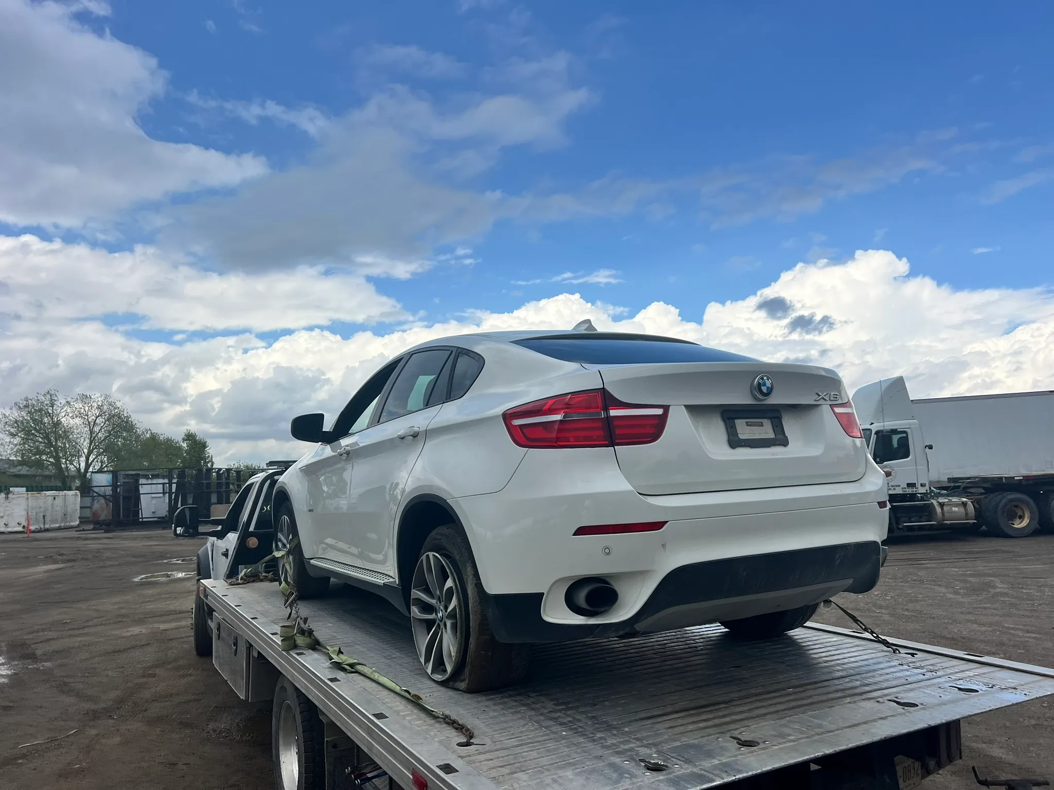 White BMW X6 SUV secured on flatbed tow truck in industrial lot; surrounded by trucks under partly cloudy sky with blue patches.