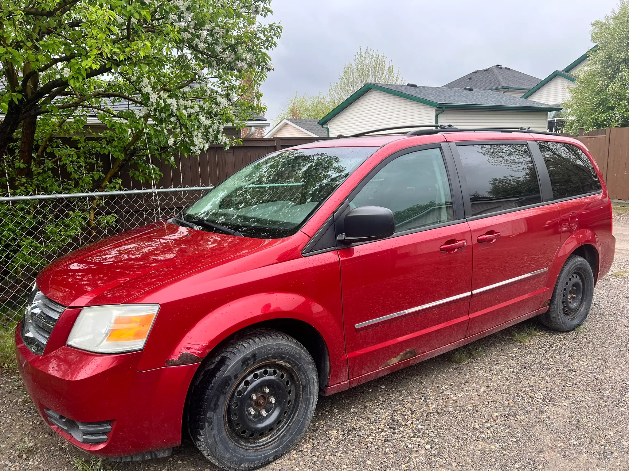 Red Dodge Grand Caravan with black steel wheels parked on gravel beside chain-link fence and leafy tree; suburban homes and wooden fence in background.