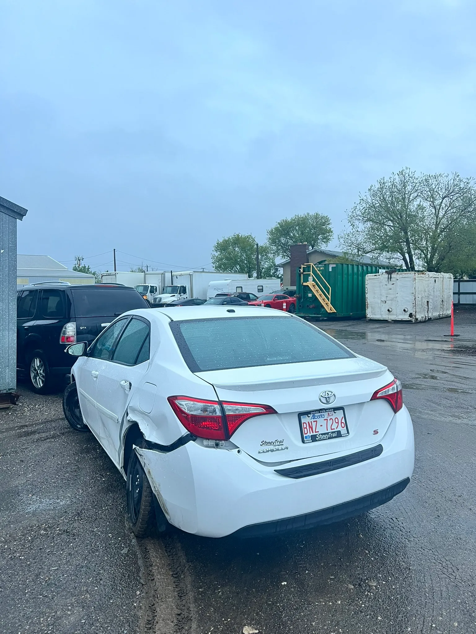 White Toyota sedan with bent rear left wheel and crumpled fender parked in lot with other vehicles and industrial containers; wet ground and raindrops suggest overcast weather.