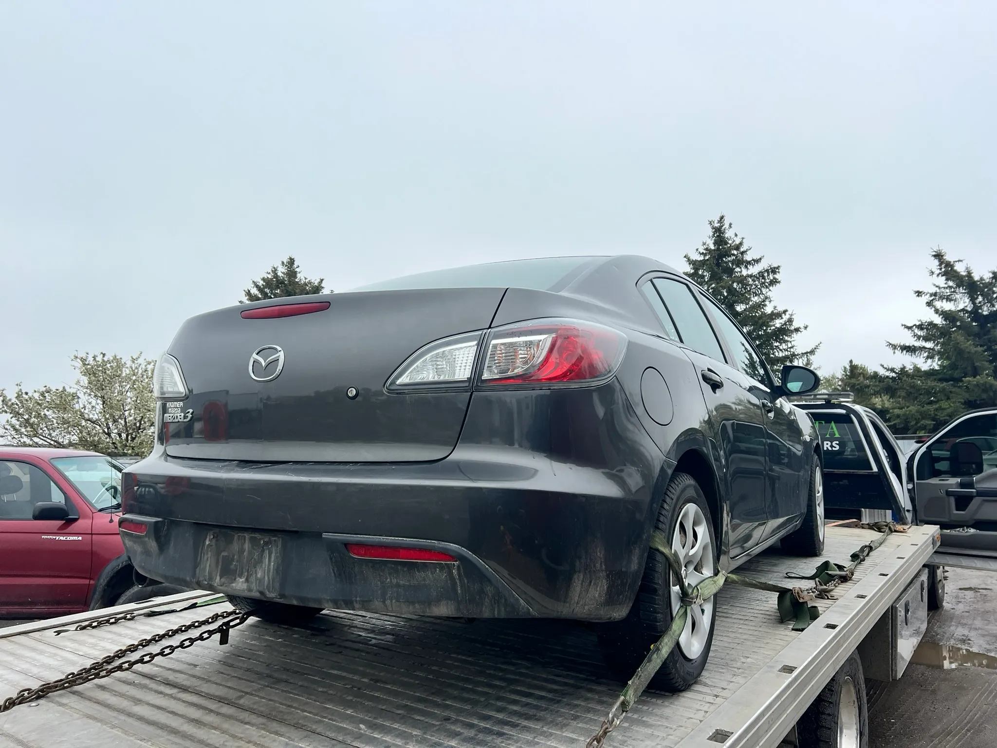 Dark-colored Mazda sedan secured with chains and straps on flatbed tow truck; parked in lot with other vehicles and trees under partly cloudy sky.