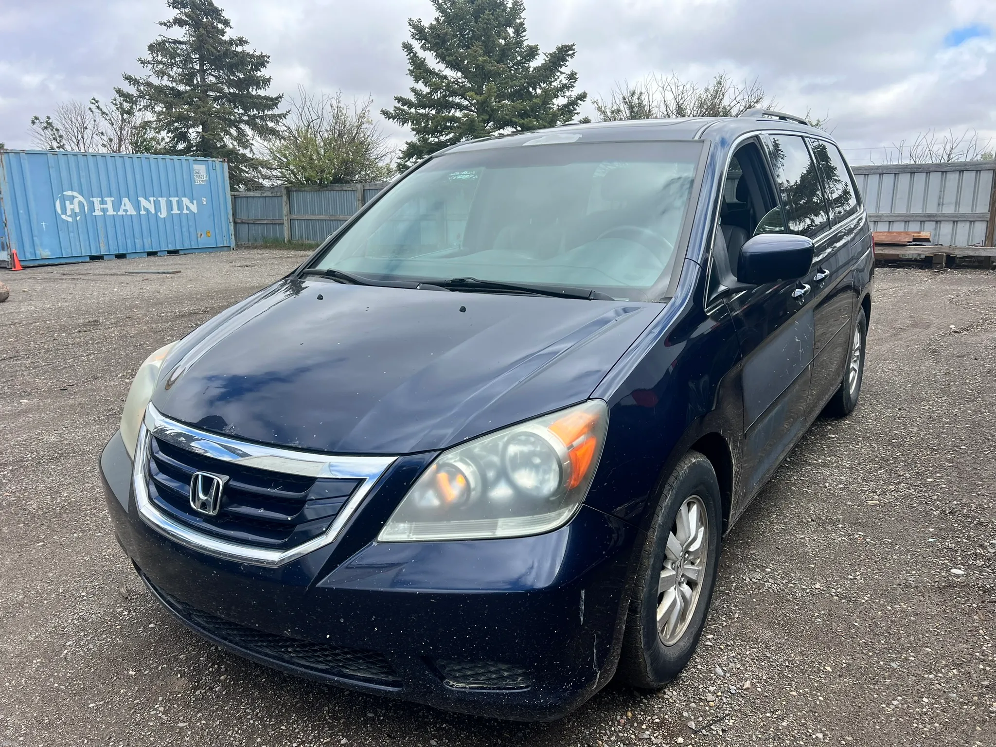 Dark blue Honda Odyssey minivan with illuminated left headlight parked on gravel near shipping containers and wooden fence; trees and partly cloudy sky in background.
