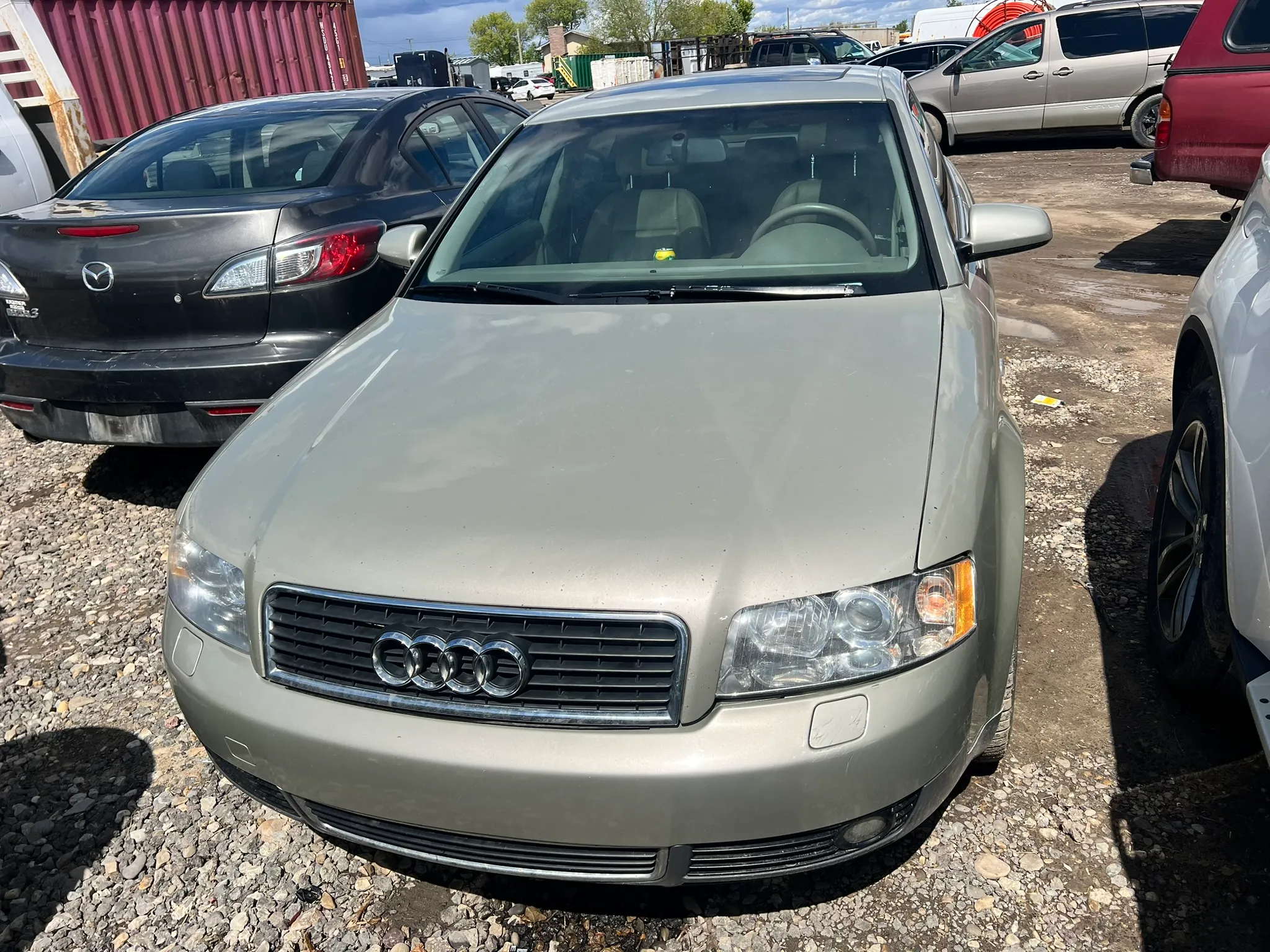 Silver Audi sedan facing forward on gravel lot among worn vehicles; industrial containers and background cars suggest salvage or resale setting.