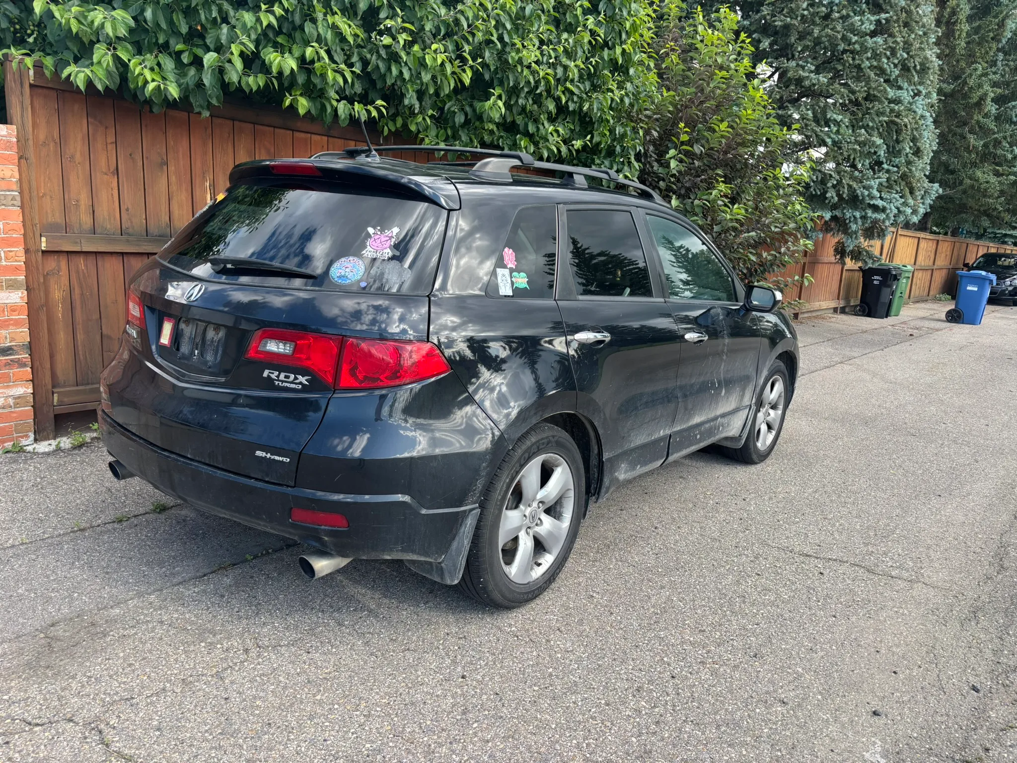 Black Acura RDX SUV with cartoon and decorative stickers on rear and side windows parked beside wooden fence; suburban street with evergreen trees and trash bins under cloudy sky.