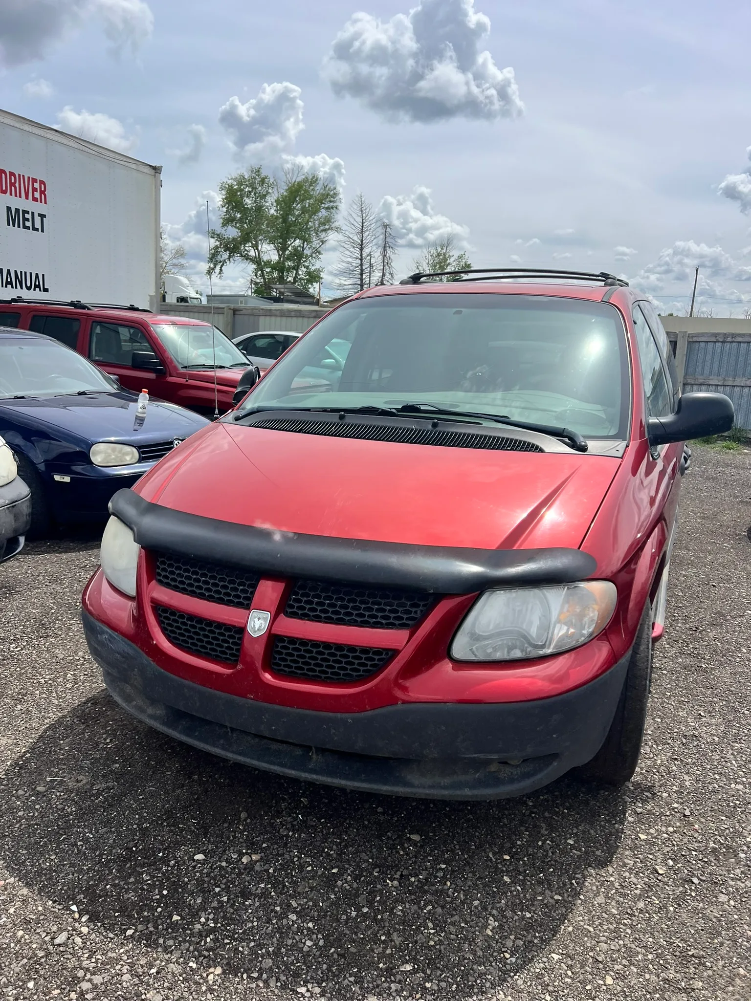 Red Dodge minivan with black hood protector and fogged headlights parked on gravel lot; white trailer with text and other vehicles in background under partly cloudy sky.