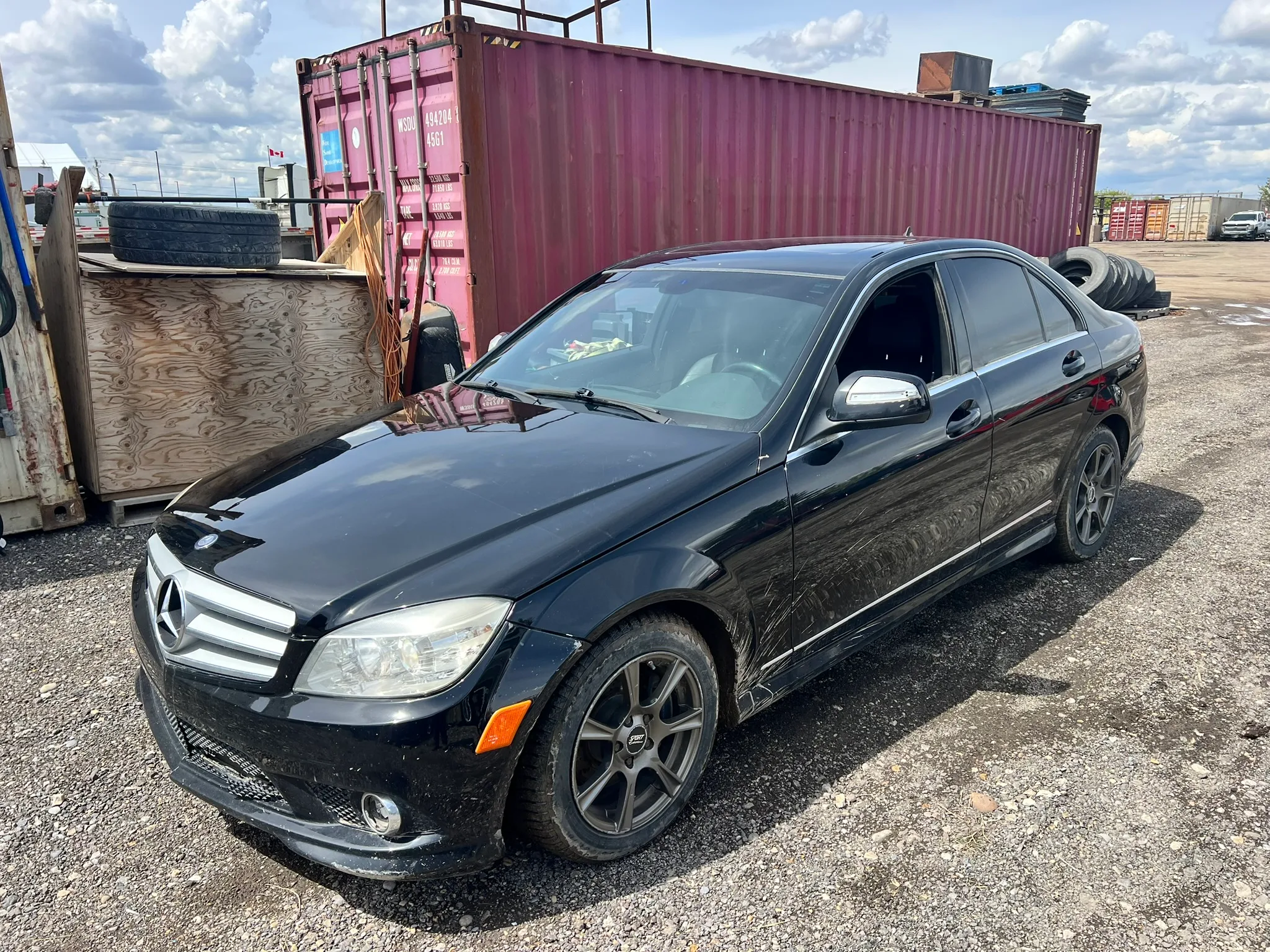 Black Mercedes-Benz C-Class sedan with tinted windows and dark alloy wheels parked on gravel in industrial yard; shipping containers, pallets, and tires in background under partly cloudy sky.